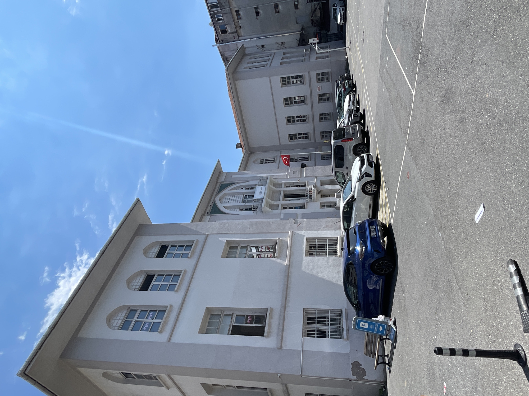 The image depicts a bright, sunny day with a clear blue sky. The scene is an outdoor area adjacent to a multi-story building with white walls and arched windows. Several cars are parked in a lot in front of the building, including a blue hatchback and a silver minivan. A Turkish flag is visible flying from a pole on the building. The ground is asphalt, marked with white lines, suggesting it could be a parking lot or a sports court. A park bench and a parking sign are also present. The overall impression is one of an institutional setting, possibly a school or a public building, located in Kadıköy, Turkey.