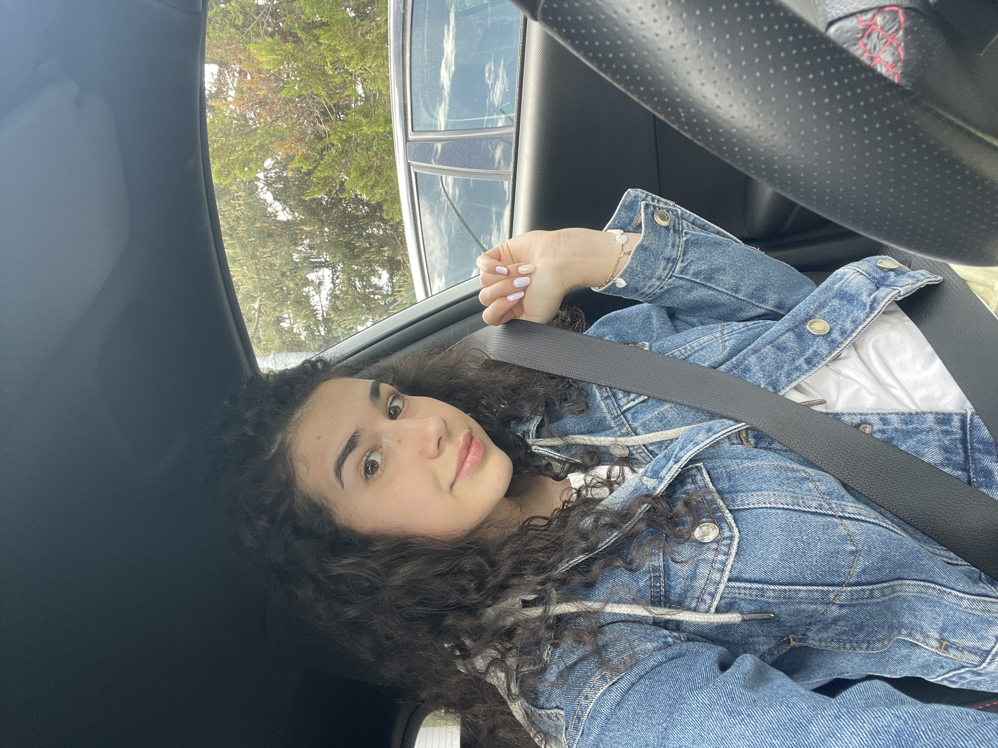 A young woman with curly dark hair is inside a car, wearing a blue denim jacket over a white hooded sweatshirt and a seatbelt. She is looking at the camera with a neutral expression. Outside the car window, trees and another car are visible. The image provides no evidence to place the location in Sofia, Bulgaria.