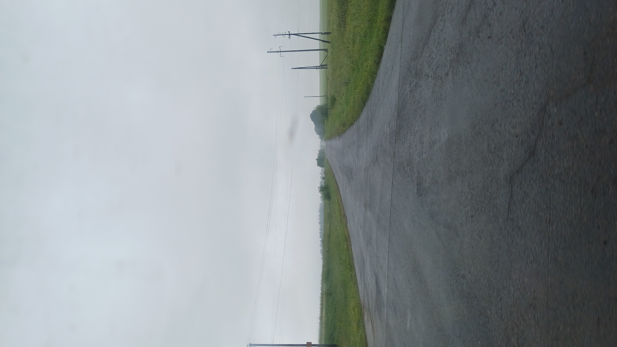 The image depicts a wet, paved road stretching into the distance through a rural landscape under an overcast sky. The road surface appears slightly reflective, suggesting recent rainfall. On either side of the road are grassy fields dotted with a few small trees and bushes. Several utility poles with power lines are visible along the road, particularly on the right side, and one on the left. The horizon is hazy, with a low-lying cloud or mist visible in the far distance. The overall atmosphere is somber and quiet, indicative of a cloudy or rainy day in a sparsely populated area. The scene is consistent with a location in Nizhnee Kechevo, Russia, given its rural setting and characteristic landscape. There are no people or vehicles visible in the image.