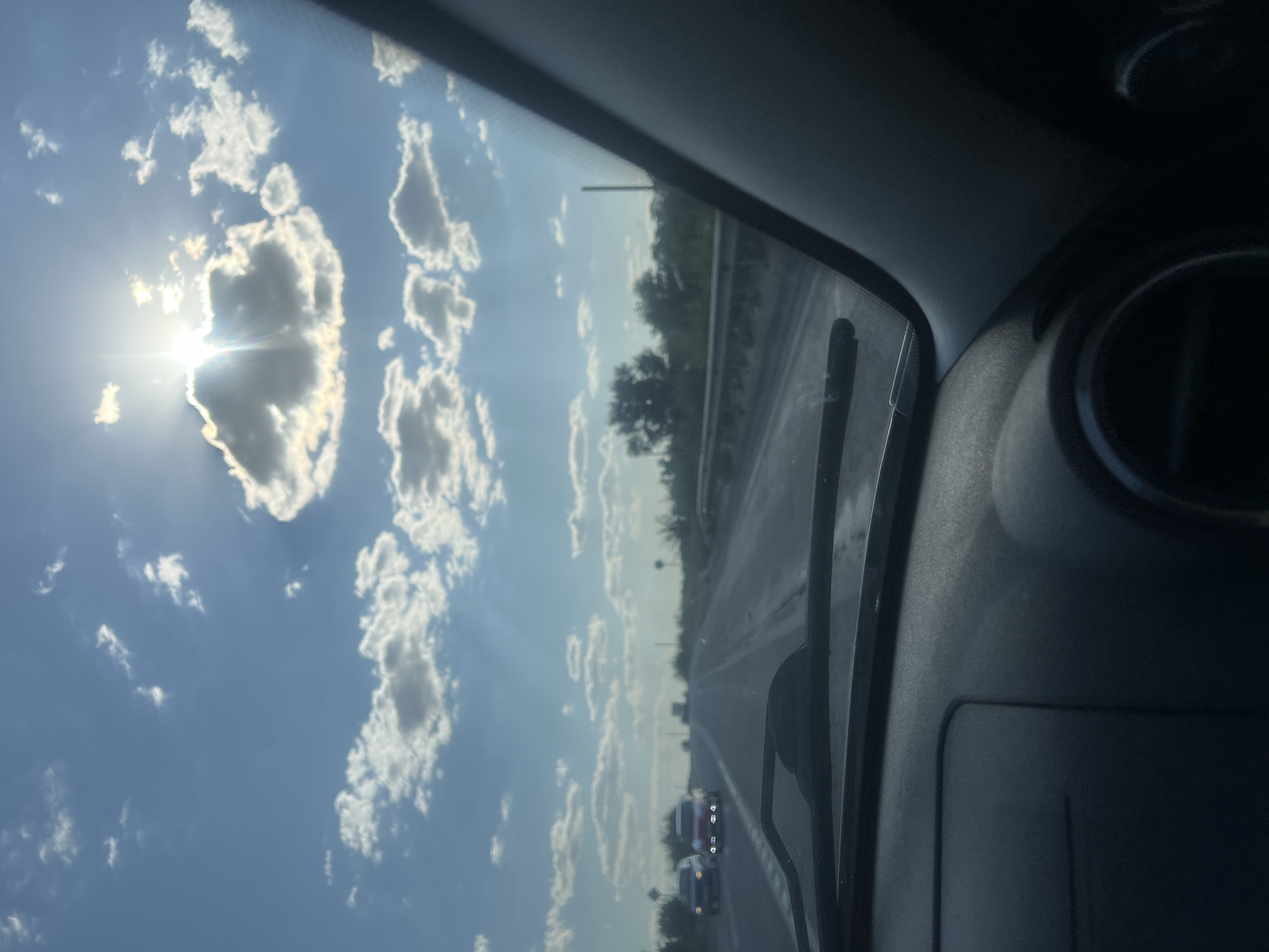 The image captures a view from inside a vehicle traveling on a highway under a partly cloudy sky with the sun partially obscured by clouds. The sun's rays are visible, creating a lens flare effect. The road ahead is visible, with a few cars in the distance and sparse vegetation and trees lining the sides of the highway. The interior of the car, including the dashboard and windshield wipers, is also visible in the foreground. The scene depicts a typical daytime highway journey in an unspecified location.