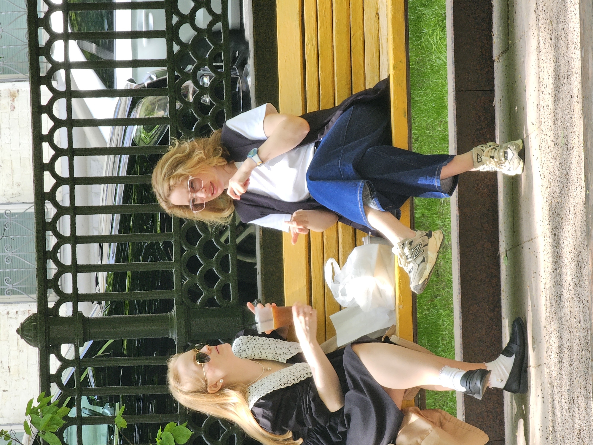 Two young women are sitting on a yellow bench in what appears to be a park in Moscow, Russia. The woman on the right is wearing a white t-shirt, a black vest, and blue jeans. She also has glasses and is wearing sneakers. The woman on the left is wearing a black dress with a white lace collar and black loafers with white socks. She is holding a drink and has a handbag beside her. There is a white plastic bag on the bench between them. Behind the bench is a black wrought-iron fence. A building and a car can be seen through the fence. The bench is on a stone or concrete surface, with grass and greenery visible around it. The scene is brightly lit, suggesting it is daytime.