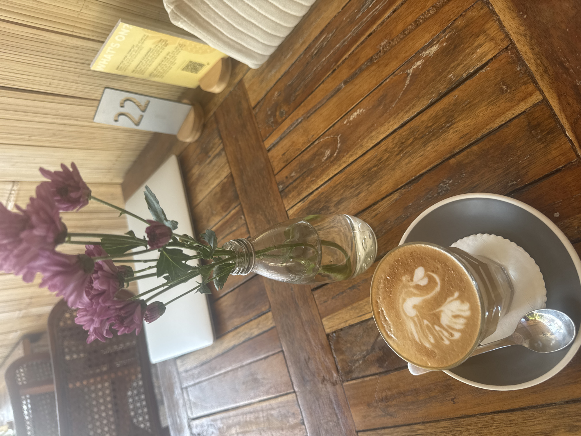 A top-down, angled shot displays a wooden table at a cafe in Nusa Penida, Indonesia. On the table, a glass of coffee with latte art resembling a swan sits on a dark gray saucer with a white lace doily. A silver spoon rests on the saucer. To the left, a small glass bottle holds a sprig of purple flowers and green leaves. In the background, a white cushion is visible, and a menu card with the words "WHAT'S ON?" printed in yellow and a QR code is propped up next to a small gray sign displaying the number "22" in a stylized font. The setting appears to be indoors or under a covered area, given the absence of direct sunlight and the presence of the cushion. The overall impression is one of a peaceful and pleasant cafe experience.