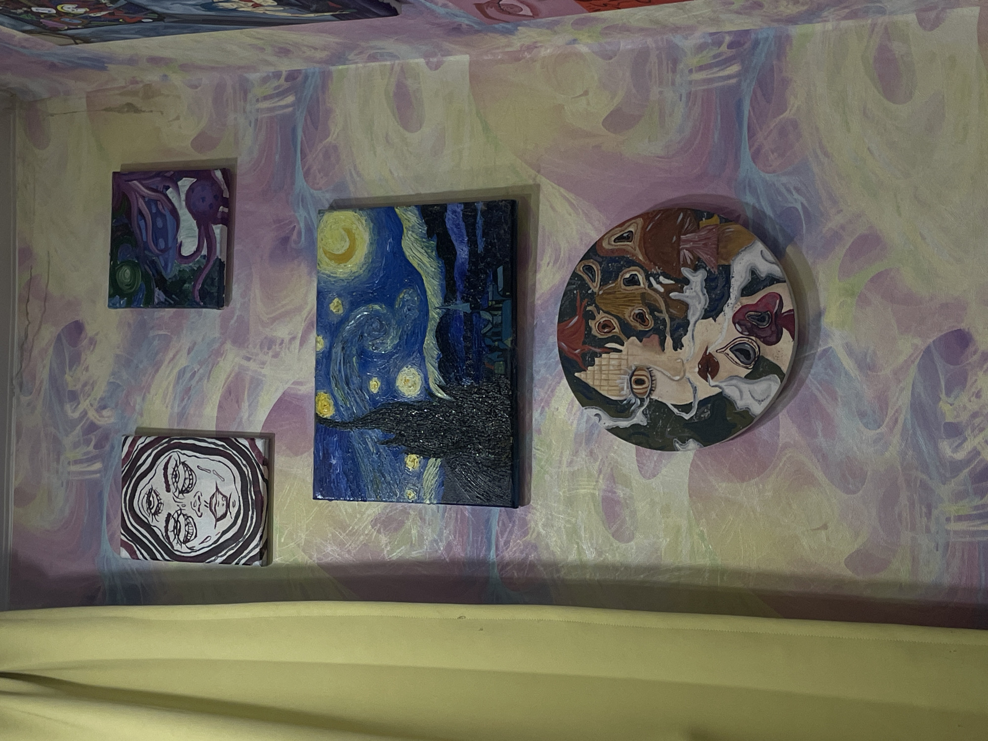 The image displays a wall adorned with several paintings. On the left, a light yellow curtain hangs. The wall itself features a psychedelic wallpaper with swirling patterns in shades of pink, yellow, blue, and purple.
There are four visible paintings:
1. **Top Left:** A rectangular painting depicting a face with three eyes, rendered in black and white with wavy lines.
2. **Top Right:** A small square painting with a purple and green abstract design, possibly representing a creature with tentacles.
3. **Middle:** A rectangular painting that is a replica of Vincent van Gogh's "The Starry Night," featuring a dark, textured cypress tree against a vibrant blue sky with yellow stars and a crescent moon.
4. **Bottom:** A circular painting with a collage-like style, featuring a face with wide, discolored eyes, red lips, and elements of mushrooms incorporated into the design.
The image appears to be taken indoors, likely in a residential setting given the presence of the curtain and the collection of artwork. There are no people or explicit actions depicted. The location is not discernible from the image itself; the provided city name, ЖК «Борисоглебское», Russia, is external information not visually present.