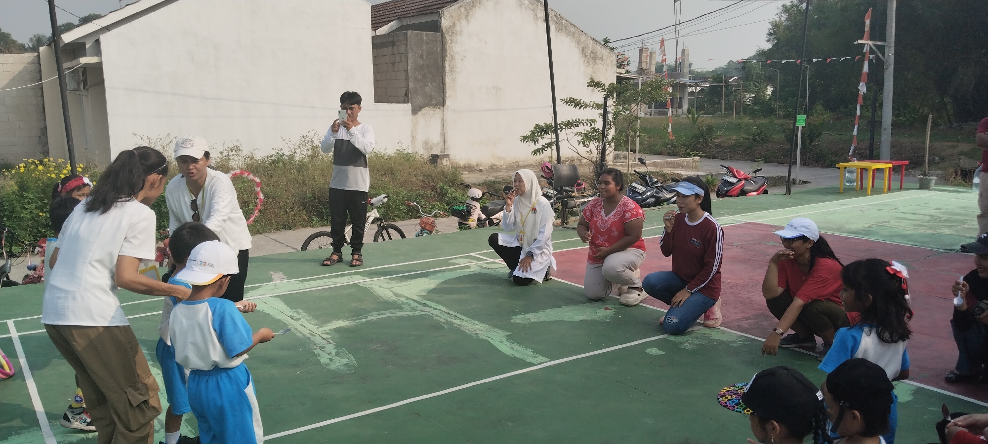 The image depicts an outdoor scene at a public badminton court, likely in Taban, Indonesia. A group of people, including adults and children, are gathered on the court. One adult male, wearing a long-sleeved shirt and black pants, stands in the middle distance holding a mobile phone. Several women and children are positioned on the court, some squatting or kneeling. A woman wearing a white shirt and cap is interacting with a child wearing a blue and white uniform, who is holding a spoon. Another child, also in a similar uniform, is standing next to them. In the background, there are residential buildings, trees, and parked motorcycles. The court itself is marked with white lines, and there are some yellow and red tables set up on the side. The overall atmosphere suggests a community event or activity taking place.