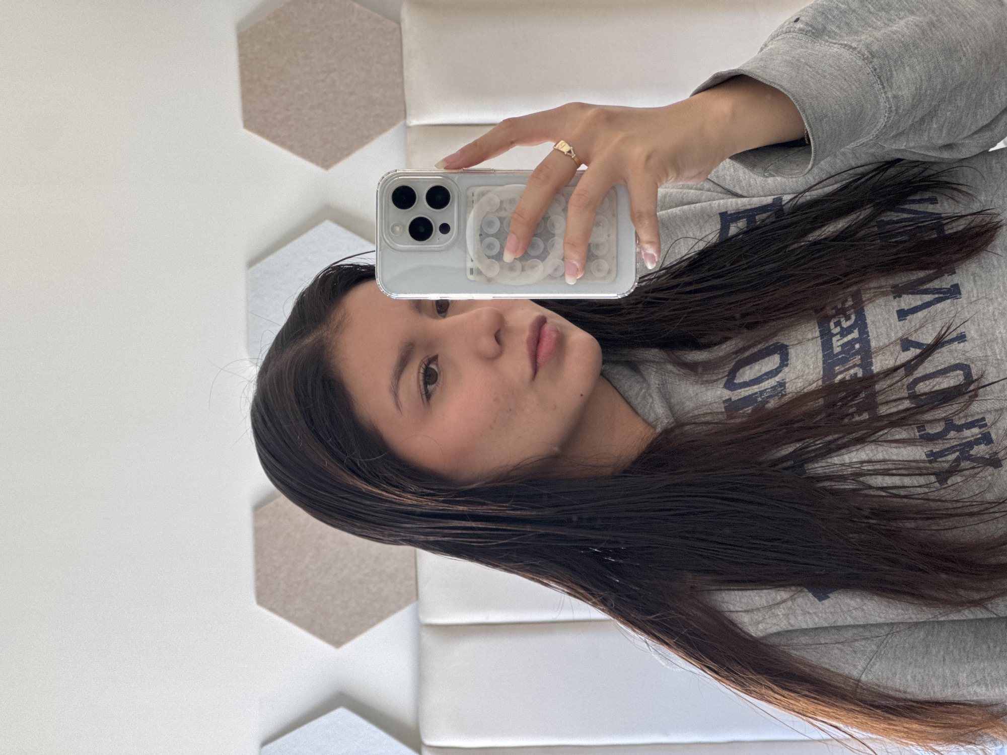A woman with long, dark hair is holding a white smartphone with a clear case, taking a selfie. She is wearing a grey sweatshirt. She has a ring on her finger. The background is a white wall with geometric hexagonal tiles in shades of grey and beige. The location is unknown.