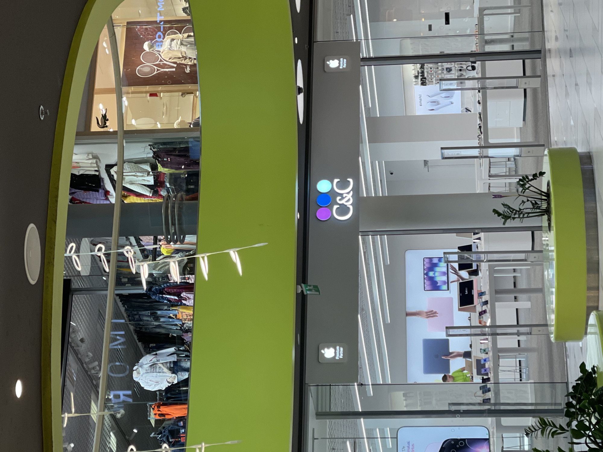 The image shows the interior of a shopping mall, likely in Tallinn, Estonia. In the foreground, a bright green, circular planter with foliage is visible. Behind it, a shop window displays various electronic devices, including laptops and phones, on white tables. The store's signage reads "C&C" with colorful circular dots above it, and an "Apple Premium Partner" logo is displayed to the right. In the background, through a glass facade, another store is visible with mannequins dressed in clothing. The upper level of the mall features more retail spaces, including one with a Lacoste logo.