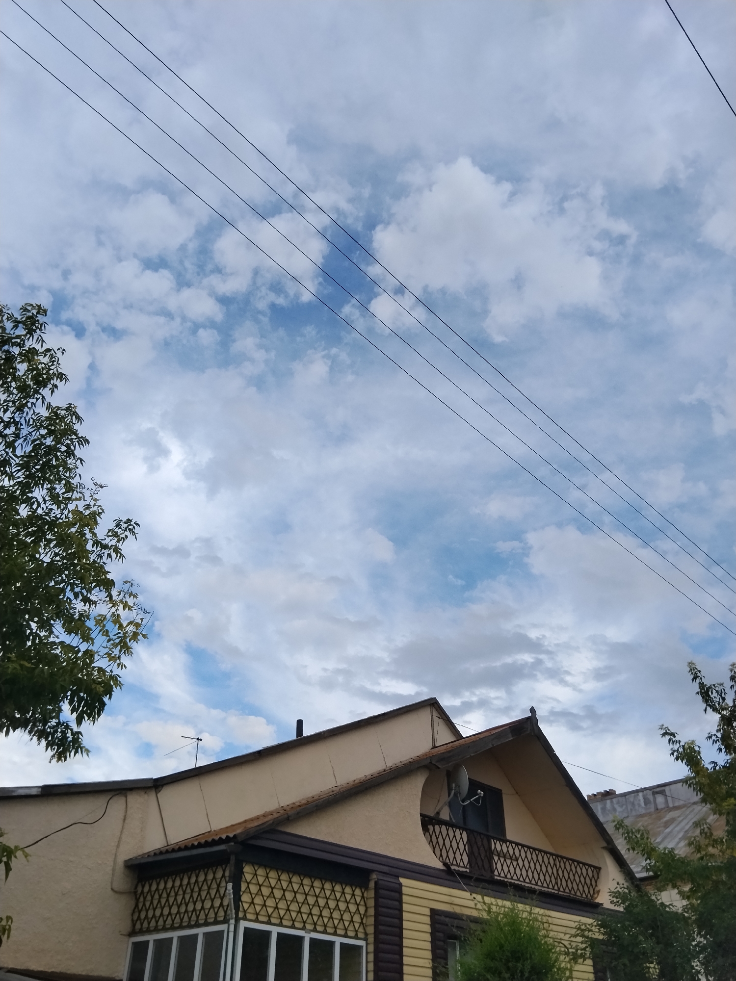 A view of a building's upper floors and roof against a cloudy sky. Four parallel power lines stretch diagonally across the frame from the upper left towards the right. A tree with green leaves is visible on the left side of the image. The building has a light-colored stucco exterior on its upper section and a yellow paneled lower section. It features a peaked roof with dark trim, a small balcony with a decorative railing, and a satellite dish mounted on the wall. Another building with a corrugated metal roof is partially visible on the right. The sky is filled with white and gray clouds.