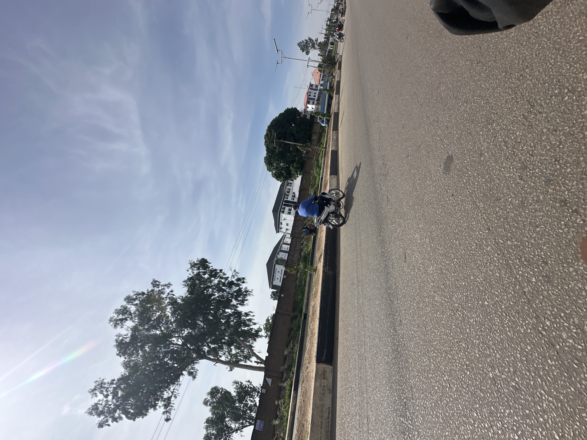 An eye-level, wide shot captures a street scene in Bukuru, Nigeria, under a partly cloudy sky. The asphalt road dominates the foreground, with a slight tilt to the left. A white motorbike with a rider in a blue shirt is traveling away from the viewer towards the right side of the frame, positioned on the road's right lane.
To the left of the road, a raised concrete curb separates the road from a strip of land with vegetation and a brick wall. Beyond the wall, several trees with lush green foliage are visible, with one large tree prominently displayed on the left. A few buildings with white exteriors and dark roofs are situated behind the trees and wall, some appearing to be residential or commercial. Streetlights with solar panels are spaced along the road in the background.
The lighting suggests it is daytime, with the sun casting a rainbow-like glare in the upper left portion of the sky, indicating a bright, potentially humid atmosphere typical of Bukuru. The overall impression is of a moderately busy urban street with elements of natural and built environments.