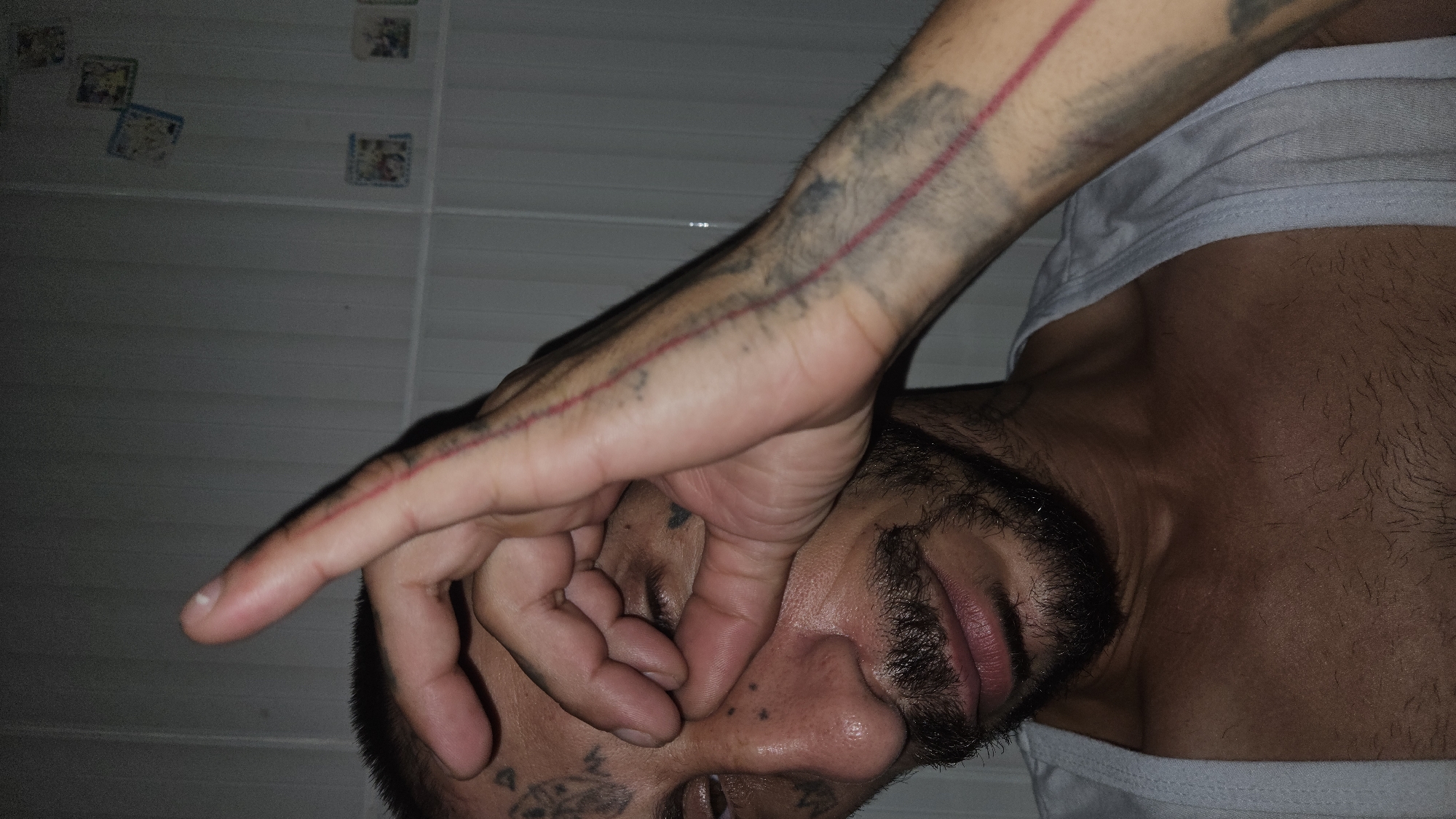 An angled, close-up shot captured in Cam Đức, Vietnam, features a person, likely male, lying down in an indoor setting. The individual's head is angled towards the bottom right of the frame, with their right hand covering their eyes and upper nose.
The person has dark hair, a beard, and prominent tattoos. A distinct red line tattoo runs along their right arm, which also displays a faded, dark, and abstract tattoo design. Additional dark, possibly symbolic or geometric, tattoos are visible on their temple or forehead area. They are wearing a plain white undershirt or tank top.
The background reveals a light-colored tiled wall, possibly suggesting a bathroom or bedroom. Several small, square decorative items, resembling stickers or framed pictures, are intermittently placed on the wall in the upper left portion of the image. The scene is dimly lit, indicating low light conditions, potentially at night. No visible text is present. The person's obscured face prevents clear interpretation of emotion, but the pose could suggest rest, sleep, or a deliberate act of covering their face.
