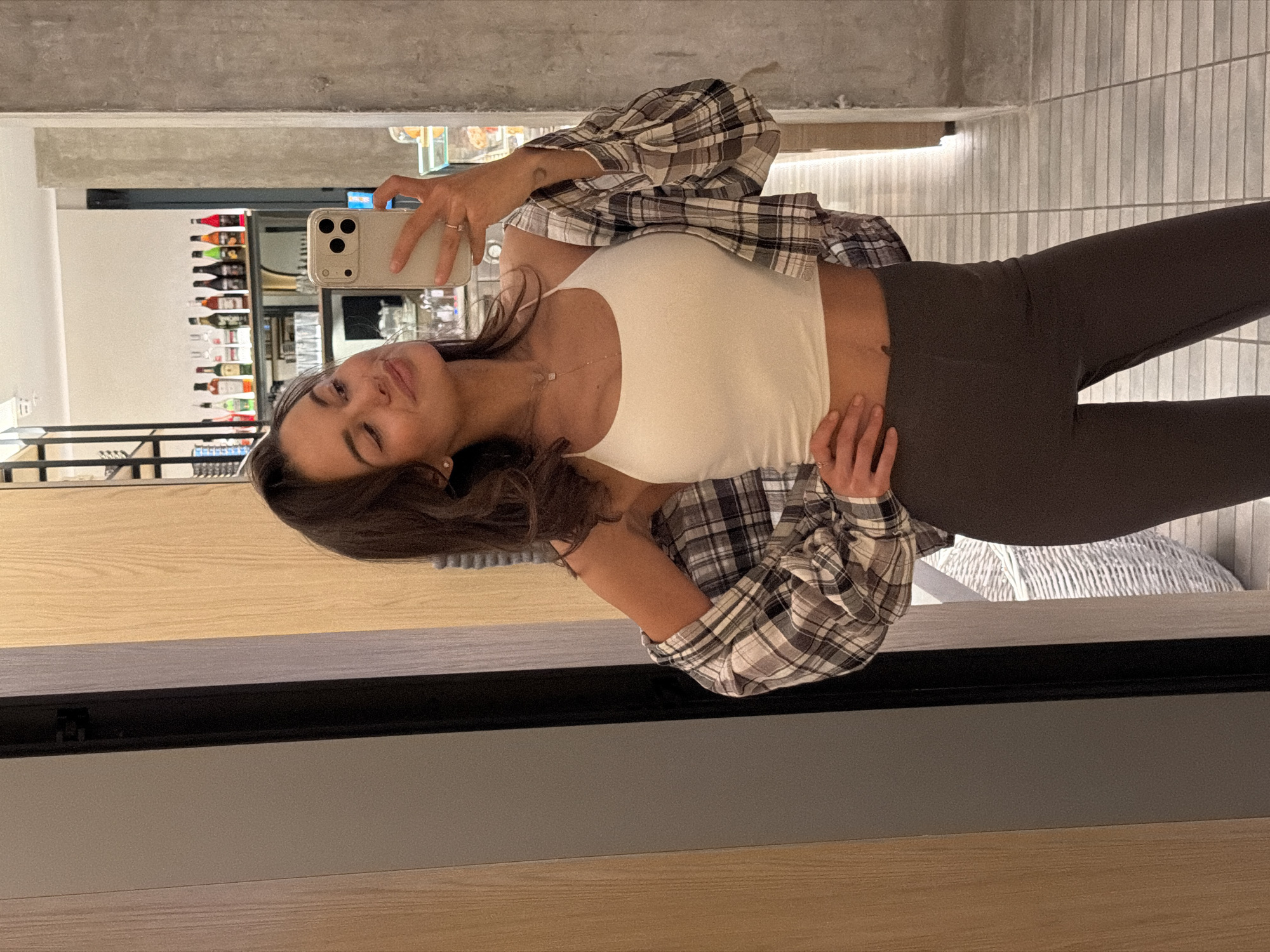 A young woman is taking a selfie in a mirror. She is wearing a white tank top and grey leggings, with a plaid shirt draped over her shoulders. She is holding a smartphone in her right hand, and her left hand is resting on her hip. The background appears to be a bar or restaurant, with shelves of alcohol bottles visible in the mirror's reflection. The location is likely in Krasnoyarsk, Russia.