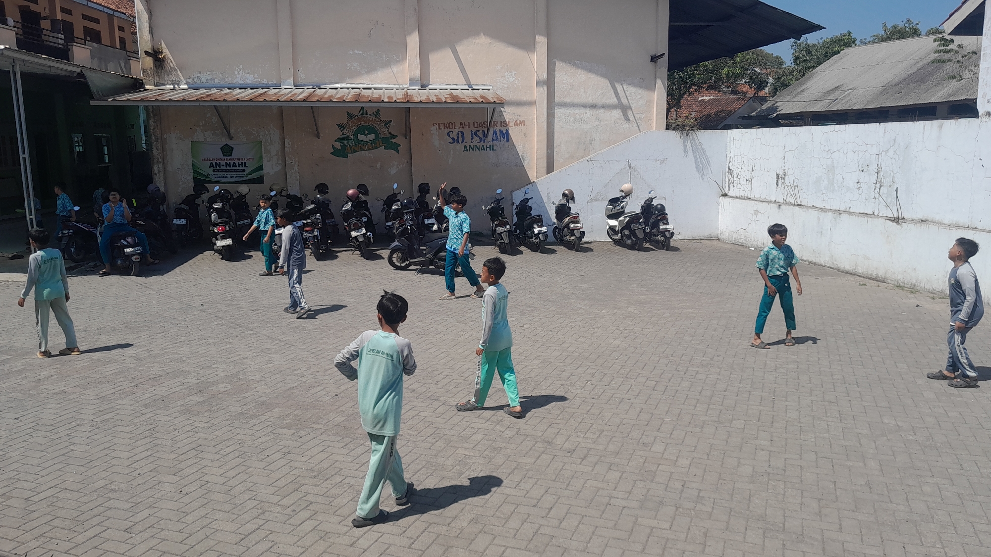 The image depicts an outdoor scene at SD Islam Ann-Nahl, located in Tasikmalaya, Indonesia. The foreground is a paved courtyard with interlocking bricks. Several children, dressed in school uniforms, are scattered throughout the scene. Two children in light blue uniforms are walking in the foreground, with one facing away from the camera. To the right, a boy in a blue and green patterned shirt and green pants stands looking upwards, with another boy in a gray and blue uniform to his right. Further back, a boy in a light blue uniform is walking towards the right side of the frame, while another boy in a similar uniform is walking past motorcycles. A group of people, including children, are seen near the building on the left, some of whom appear to be sitting or standing near parked motorcycles. The background features a light-colored building with signs and a mural depicting the school's emblem. A row of parked motorcycles is visible in front of the building. The sky is clear and blue, indicating a sunny day.