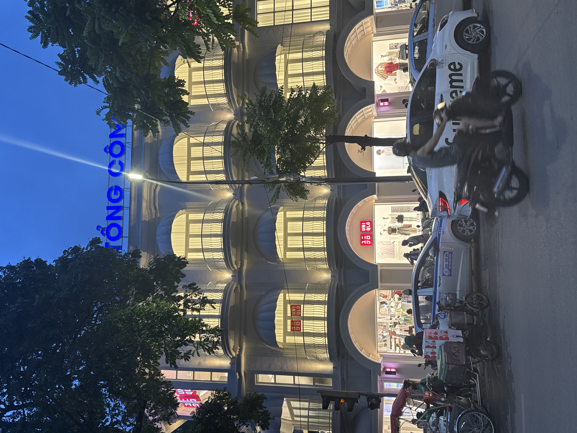 Captured at dusk in Hà Nội, Vietnam, the image showcases an architecturally prominent building illuminated against a darkening sky. The building, possibly a department store or commercial edifice, features multiple stories with arched windows, each radiating light from within. A blue, vertically-oriented sign, possibly displaying "Tràng Tiền," is visible on the left side of the building.
On the street level, cars and motorbikes are parked alongside the building. One white car parked is labeled with "supreme" on its side. A motorcyclist is captured in motion, slightly obscuring a vehicle. Other vehicles, including a bicycle-powered cart, are stationed near the sidewalk. The presence of trees and a street lamp further enriches the urban setting.