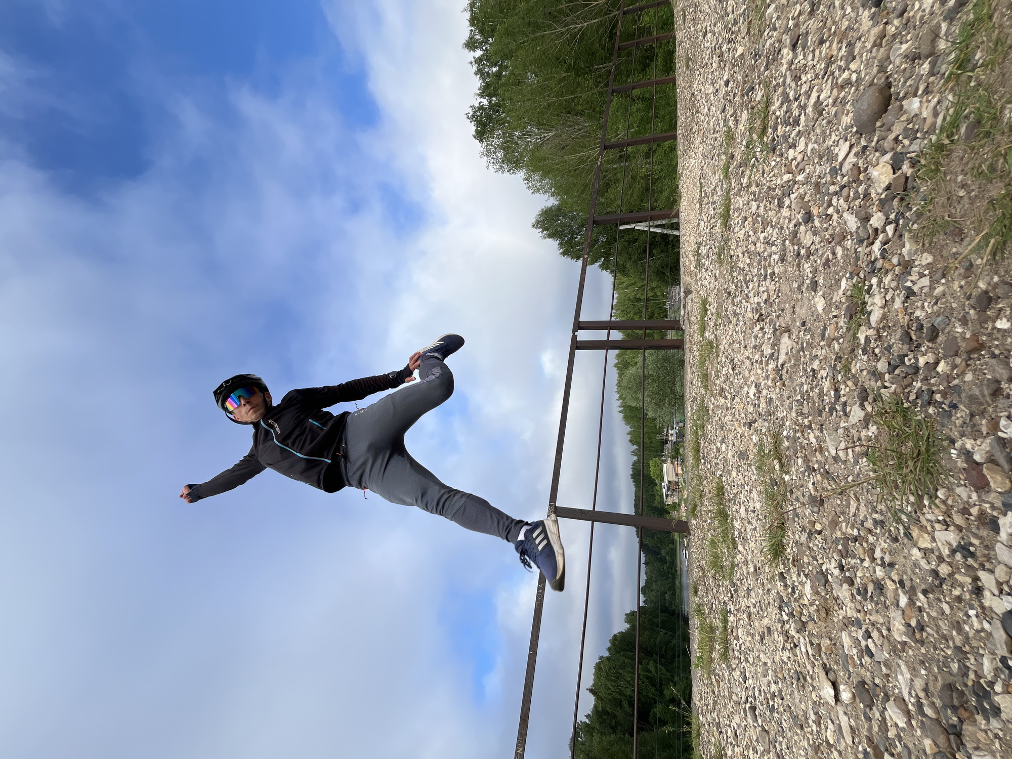 The photograph captures a low-angle, wide-shot perspective of a person in mid-air, performing a jump over a metal railing. The individual, wearing a black jacket with blue accents, grey sweatpants, athletic shoes, a helmet, and sunglasses, is positioned in the upper-left quadrant of the frame, their body angled towards the right. Their left arm is raised and their right leg is bent, appearing to propel them over the horizontal bar of the railing. The railing itself is weathered and spans the mid-section of the image horizontally.
Beyond the railing, the background depicts a lush green forest that extends to the horizon, suggesting a natural or semi-natural environment. A body of water, possibly a river or lake, is visible in the middle distance, reflecting the sky. A few small structures, appearing to be buildings, are scattered along the opposite bank of the water. The sky is a dynamic mix of blue with scattered white clouds, indicating a partly cloudy day.
The foreground is dominated by a rough, gravelly surface interspersed with sparse green grass, suggesting a path or embankment. The image appears to have been taken in Chaikovsky, Russia, given the context of the city's geography, which features the Kama River and surrounding natural landscapes. The overall impression is one of energetic movement against a backdrop of nature and the sky.