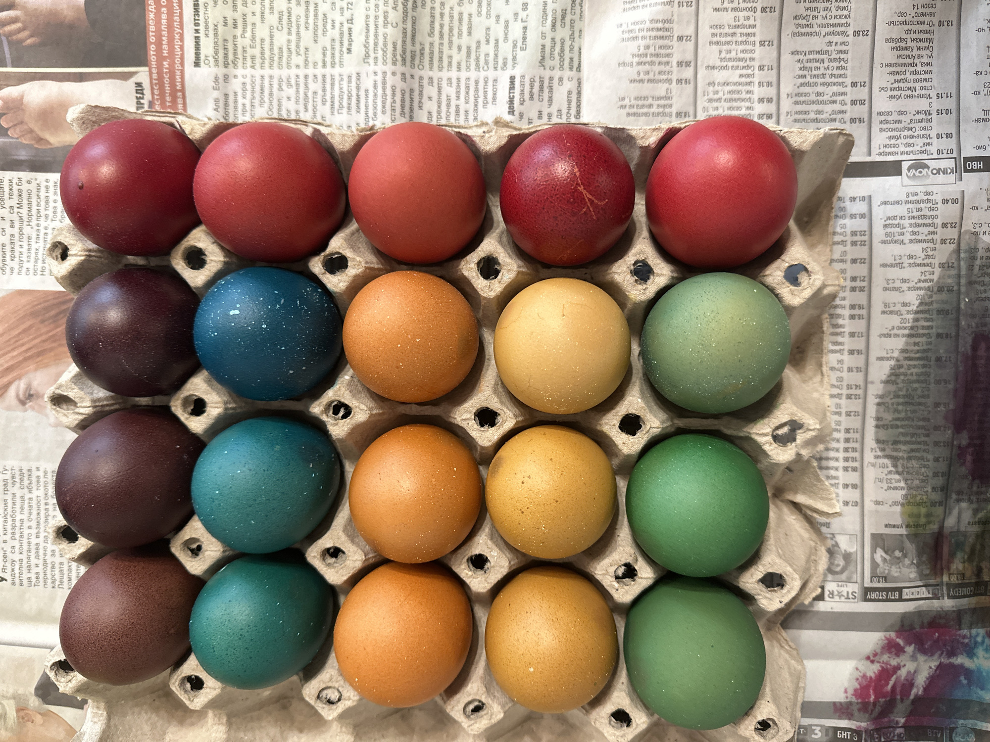 The image shows a carton of twenty Easter eggs arranged in four rows and five columns. The eggs are dyed in various colors: red, dark brown, blue, orange, yellow, and green. The carton sits on a newspaper in the Bulgarian language, suggesting the image was taken in Bulgaria. A partial view of a foot is visible in the upper left corner of the image, also on the newspaper. Newspaper articles include the words "BTV" and "KINONOVA." Based on the text "Втората световна война: цената на империята, сезон 1, еп. 13" on the newspaper, the location could be Sofia, Bulgaria, where newspapers in Bulgarian are commonly found.