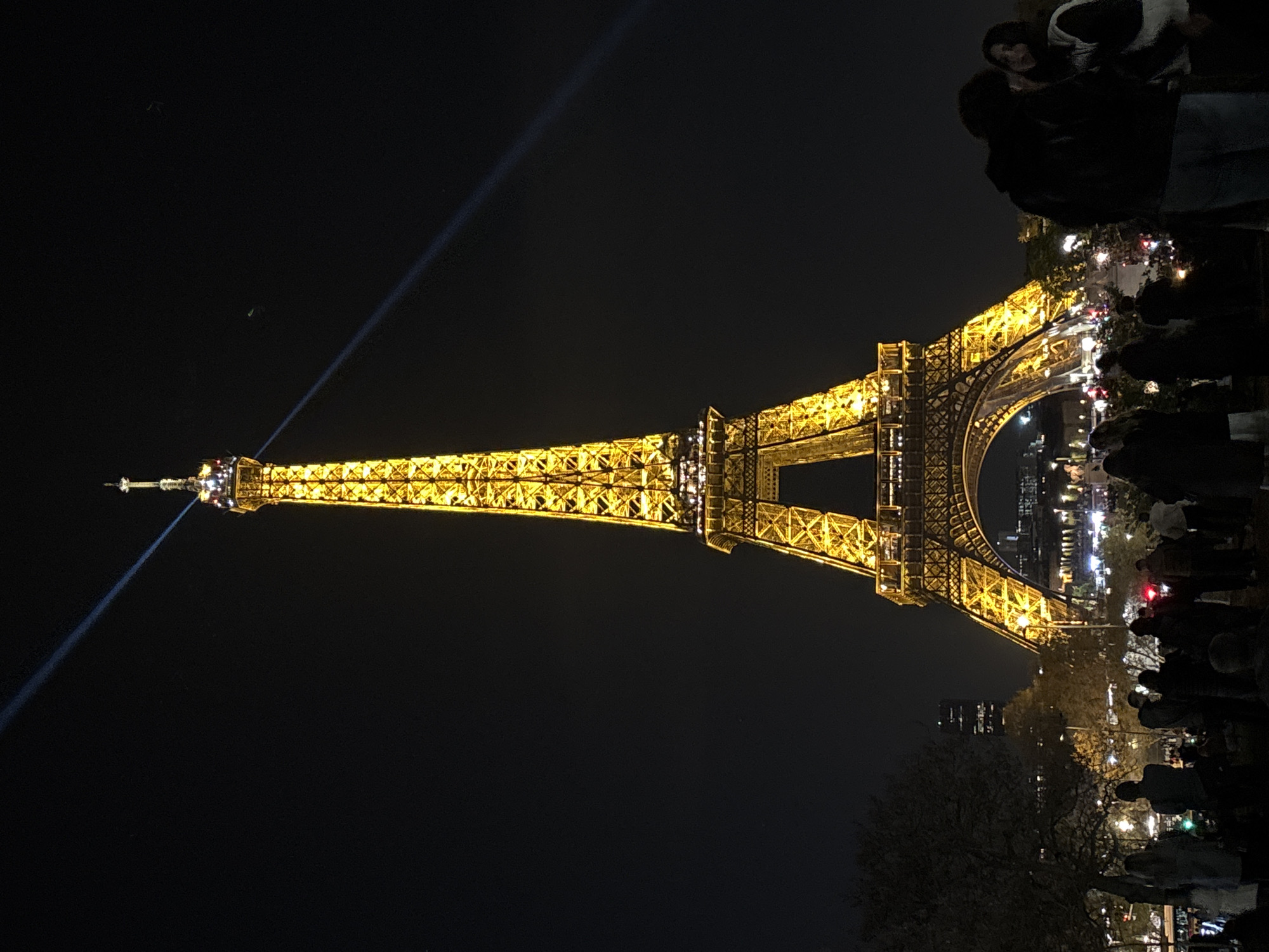 The image is a nighttime shot of the Eiffel Tower in Paris, France. The tower is illuminated with golden lights and stands prominently against the dark sky. A beam of light projects upwards from the tower. A crowd of people is gathered at the base of the tower, presumably observing and taking pictures. In the background, city lights are visible, indicating the urban environment of Paris. Some trees are also present near the crowd.