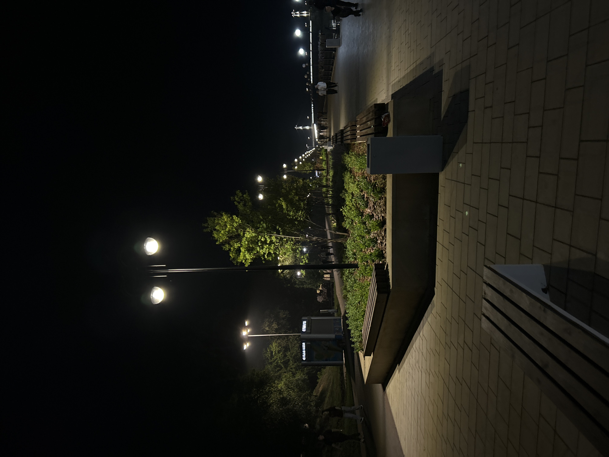 The image depicts a nighttime scene along a paved promenade, likely a public park or waterfront area in Orenburg, Russia. Several streetlights illuminate the path, casting long shadows. The promenade is lined with benches and planters containing greenery. In the background, more streetlights stretch into the distance, suggesting a long walkway or bridge. A few individuals are visible walking along the promenade, appearing as silhouettes against the illuminated path. The overall atmosphere is calm and orderly.