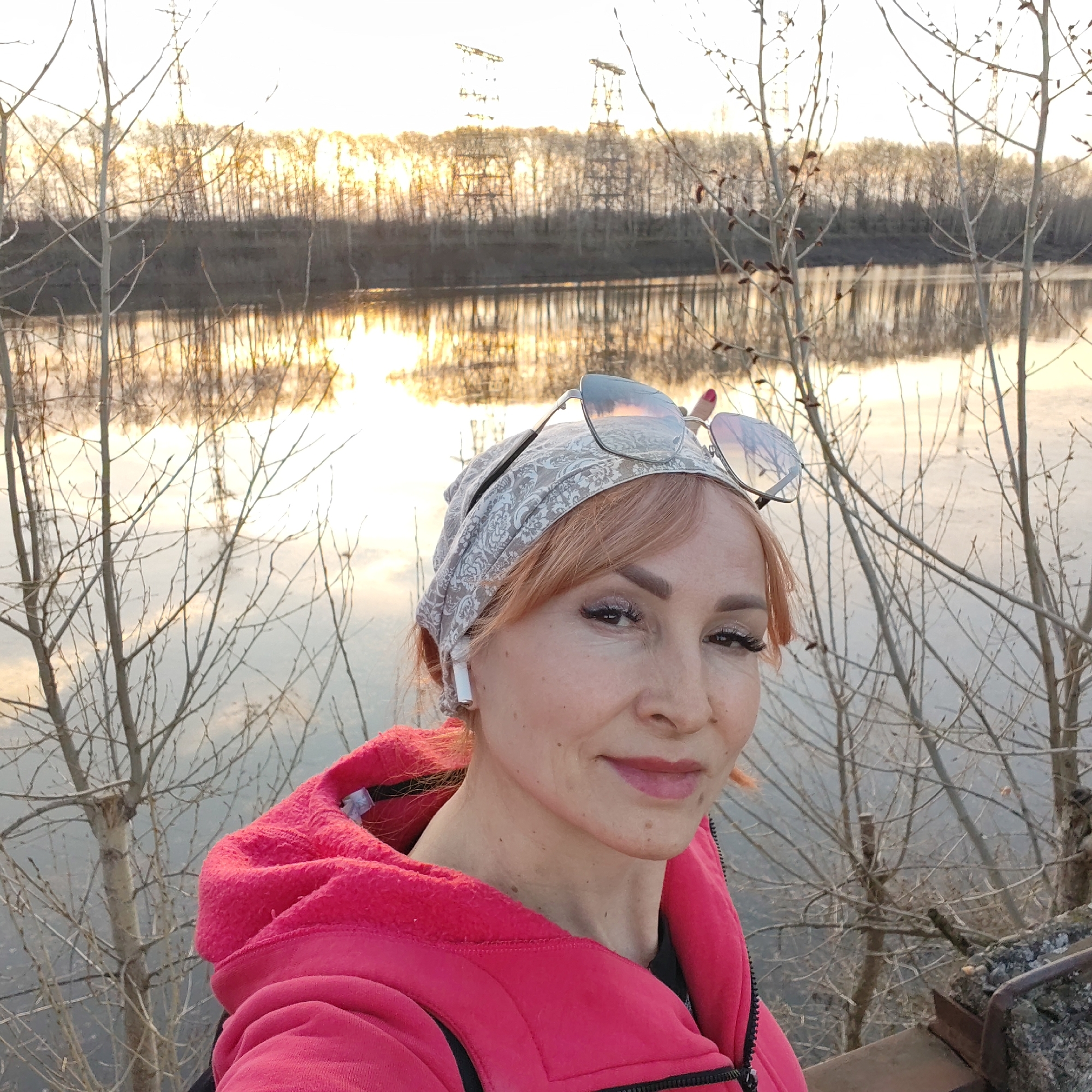 A woman with pale complexion, pink hair, and a headscarf poses in front of a river at sunset in Chaikovsky, Russia. She wears sunglasses on her head and a pink hooded sweatshirt. The river reflects the golden light of the setting sun. Bare trees are interspersed between the woman and the river. Power line towers are visible in the background.