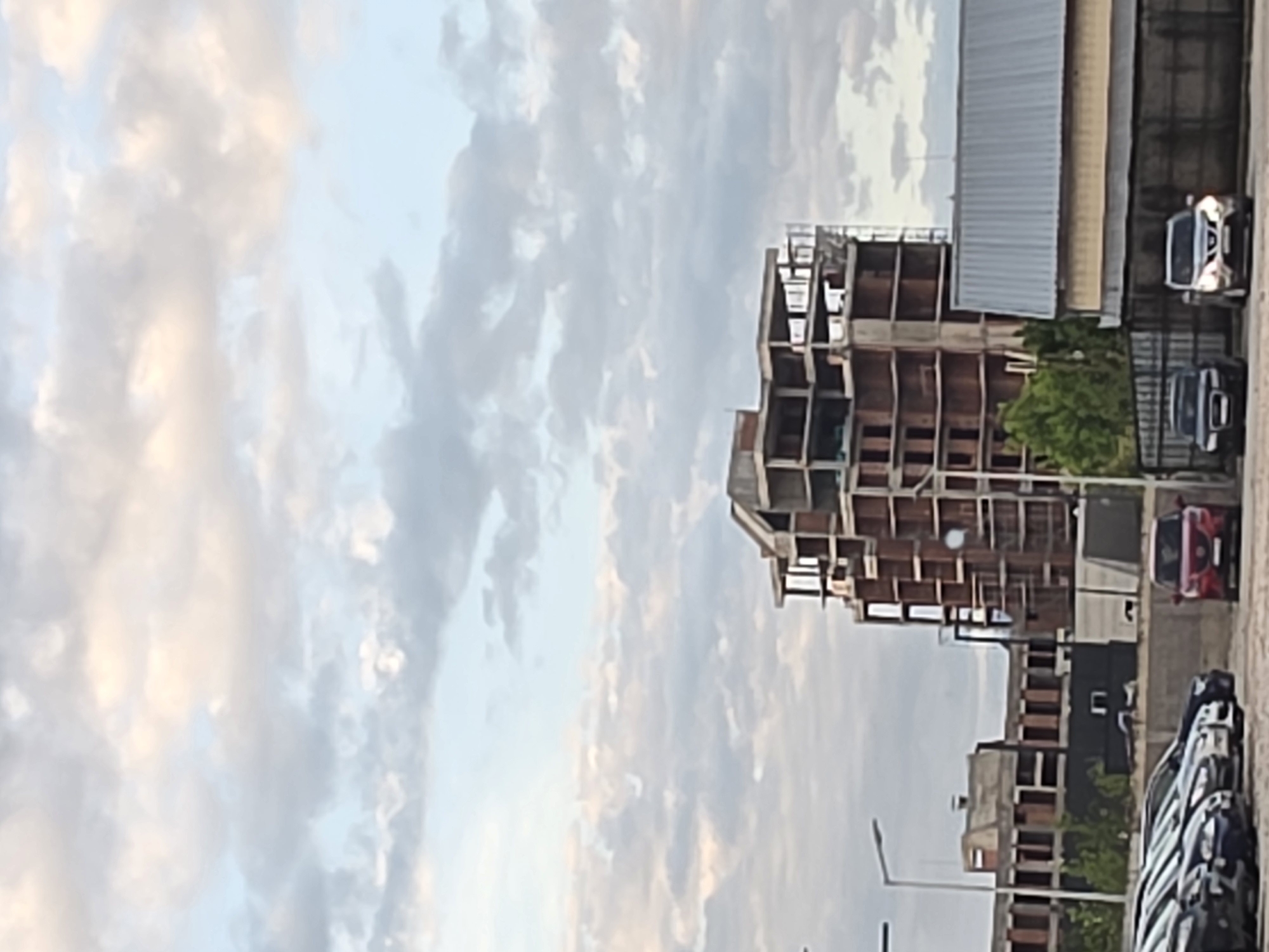 The image shows a construction site in Plovdiv, Bulgaria. A multi-story building is under construction, with the frame visible. There are cars parked nearby, and another building can be seen in the background. The sky is cloudy. There are street lights visible, and the scene appears to be taken from a street level.