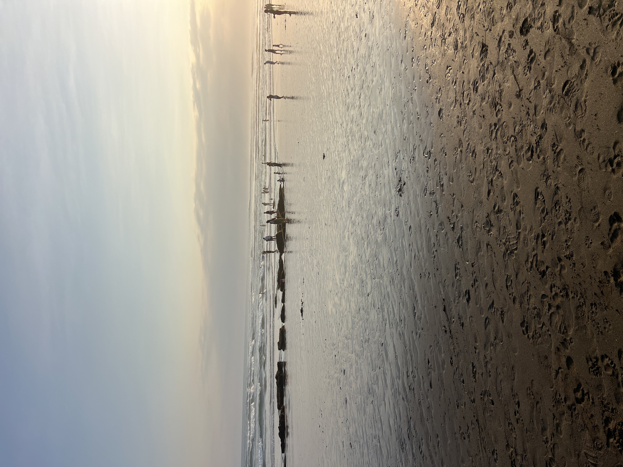 The image depicts a beach scene in North Kuta, Indonesia. The sand in the foreground is wet, reflecting the sky and appearing textured with numerous footprints. In the mid-ground, the ocean meets the shore with moderate waves breaking. A line of dark, rocky formations protrudes from the water and extends across the beach. Several individuals, appearing as silhouettes due to the backlighting, are scattered along the water's edge and on the wet sand. Some are standing, others appear to be walking or interacting with the water. The sky above is a gradient of pale blue and soft yellow, with thin clouds visible near the horizon, suggesting it may be late afternoon or early morning.