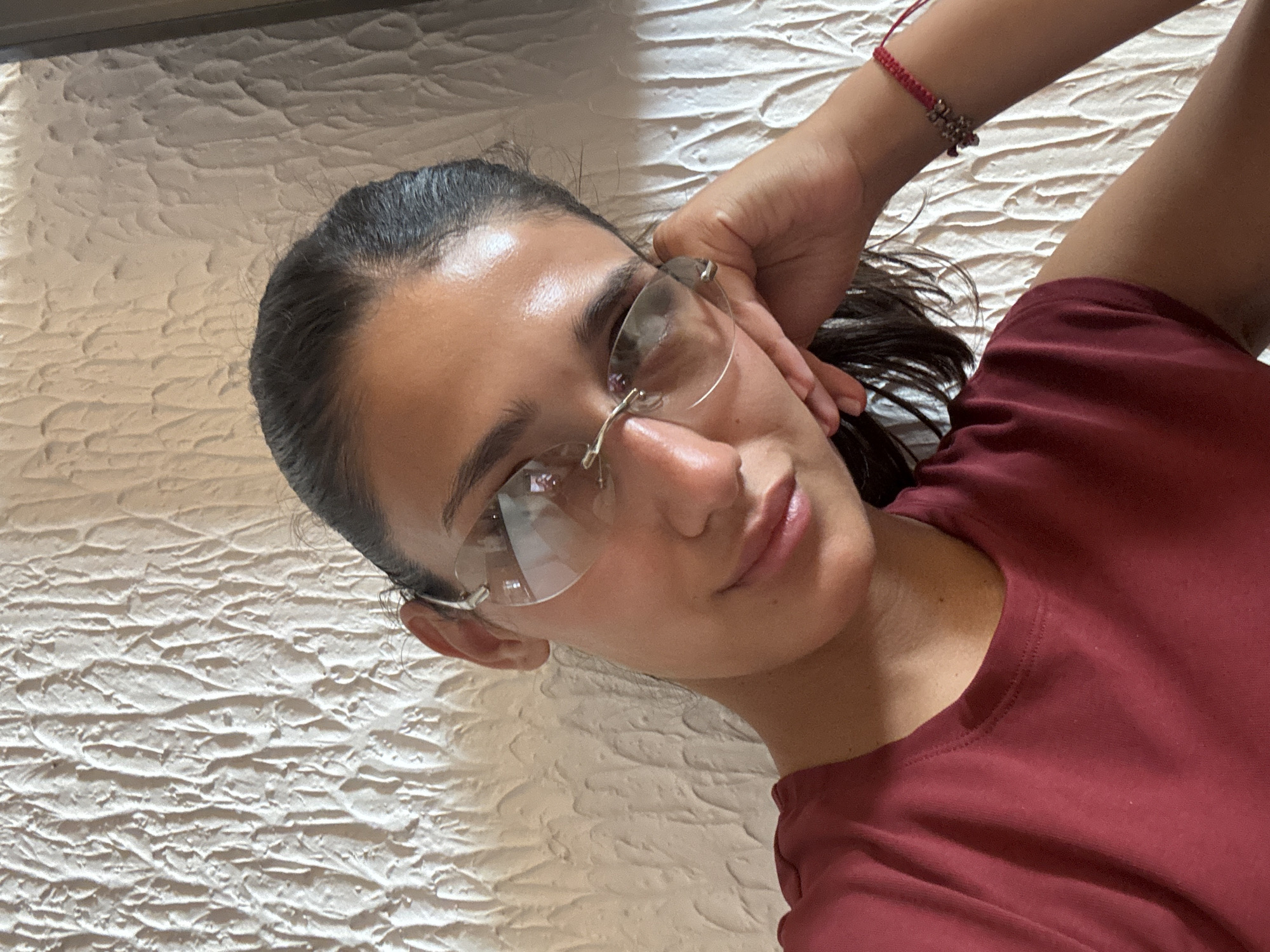 A young woman is pictured in a selfie, leaning her head on her hand, and wearing glasses. She has dark hair pulled back and is wearing a maroon t-shirt. She has a red bracelet on her right wrist. The background is a textured white wall. The image's metadata does not contain any information about the location in Miljevci, Montenegro.