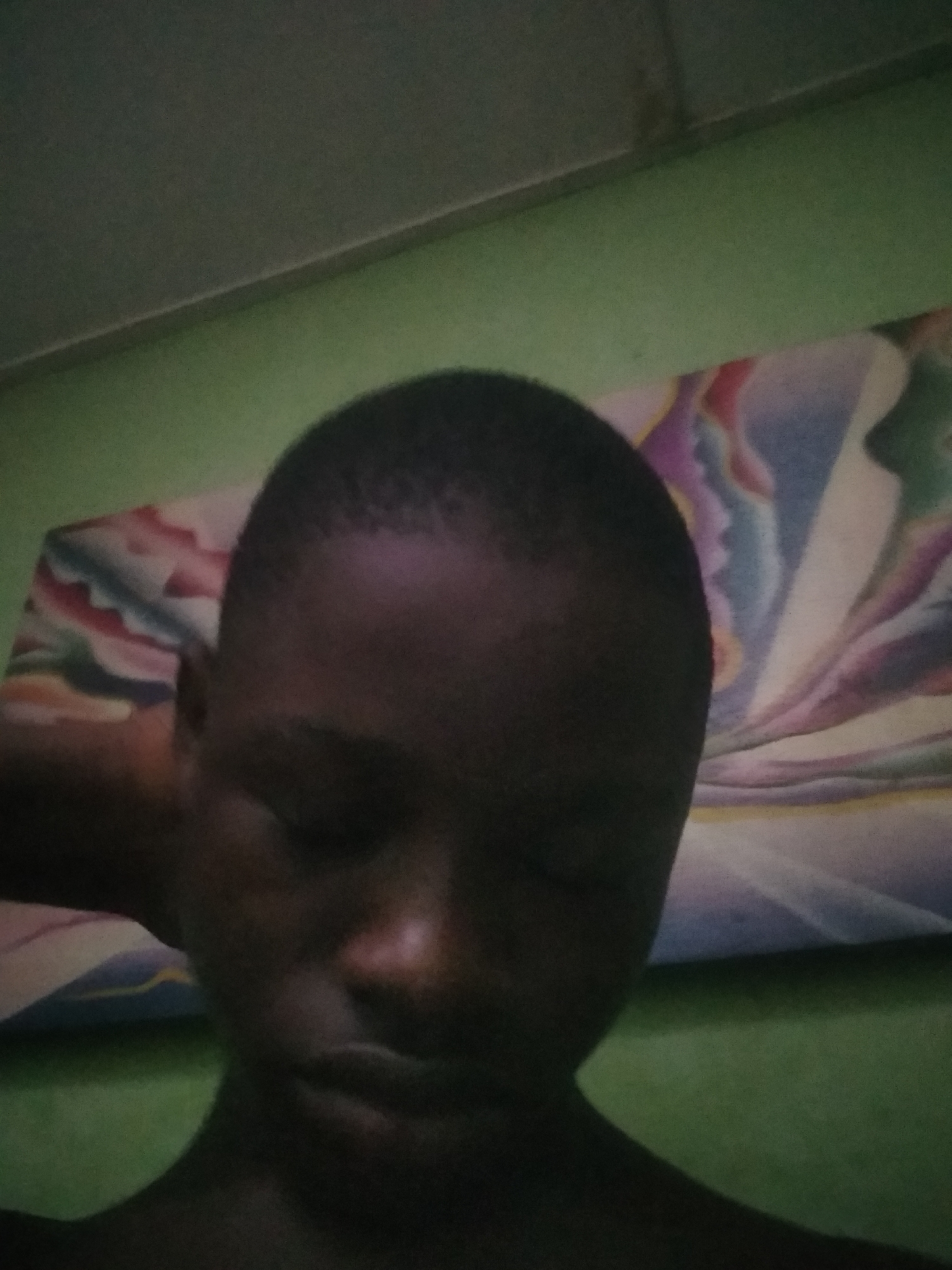 A low-light, close-up portrait shows a young person, likely male, with dark skin and short hair. Their head is tilted downwards, eyes appearing closed or looking away, suggesting a state of rest or sleep. A faint highlight catches the left side of their face, illuminating the bridge of the nose and cheek against the dim surroundings.
Behind the person's head and left shoulder, a colorful patterned object, possibly a pillow or blanket, is visible. It features abstract, wavy designs in various shades, including blue, purple, pink, white, and green. The background consists of an indoor setting with a light green wall in the lower section, topped by a darker, shadowed area, likely a ceiling or upper wall, separated by a distinct horizontal line. The overall lighting is very dim, creating a subdued atmosphere. No text is visible.