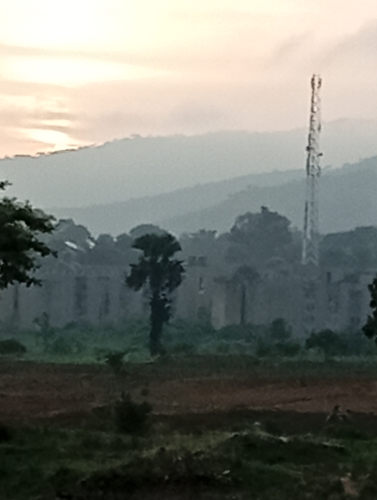 The image depicts a hazy, overcast sky over a landscape that appears to be on the outskirts of Jos, Nigeria. In the mid-ground, a tall, slender telecommunications tower stands prominently, its metallic structure faintly visible against the muted sky. Behind the tower, a line of layered hills recedes into the distance, gradually fading in clarity due to atmospheric conditions.
In the foreground, the scene transitions to a more rural or undeveloped area. A long, low structure, possibly a building or a series of interconnected structures, is visible behind a scattering of trees and vegetation. The architecture of this structure is indistinct, appearing somewhat dilapidated or incomplete, with visible openings and a weathered facade. To the left and right, the dense foliage of trees frames the mid-ground.
The immediate foreground is a darker, more textured area, likely representing cultivated land or rough terrain, with patches of green vegetation interspersed with darker, ploughed soil. The overall impression is one of a vast, somewhat undeveloped expanse under a diffused light, characteristic of early morning or late afternoon in a tropical or sub-tropical region. The presence of the telecommunications tower suggests modern infrastructure within a less urbanized setting.