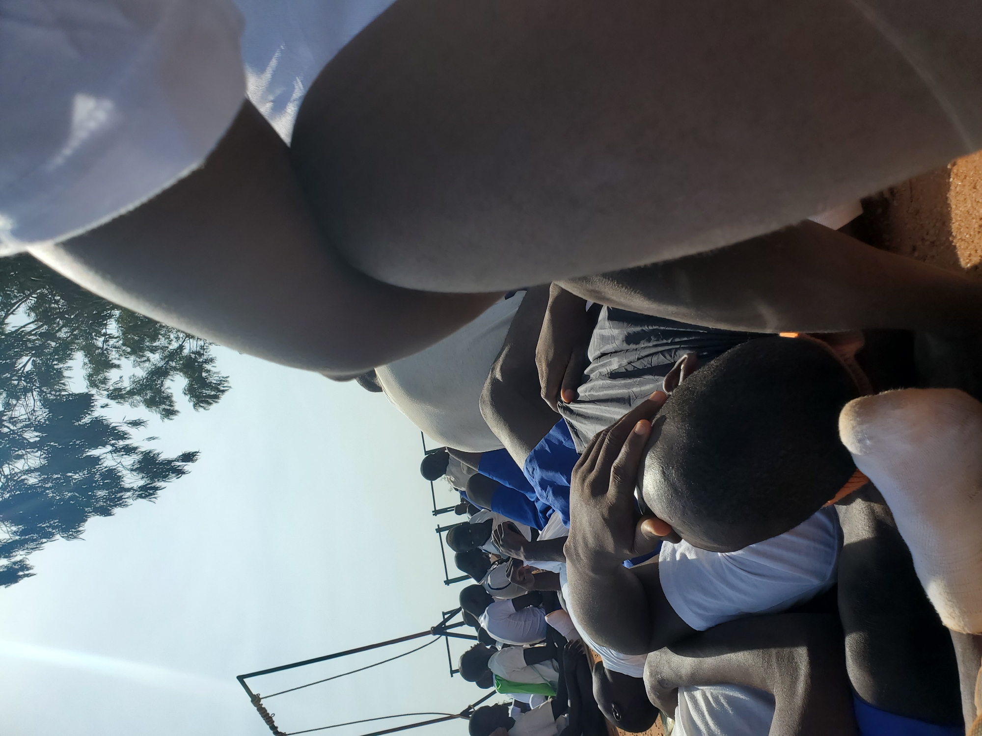 In the city of Barakin Rafin Gora, Nigeria, a group of young men, likely students based on their uniforms of white shirts and blue shorts or trousers, are seen lying on the ground outdoors. The scene is characterized by a bright, possibly hazy, sky with sunlight filtering through the leaves of a tree overhead. A metal structure, resembling part of a swing set or some form of playground equipment, is visible in the background. The individuals are resting, some with their eyes closed, in what appears to be a relaxed or resting state. Their clothing suggests a communal setting, possibly a school or youth camp. The ground is covered in sand or dirt. The focus of the foreground is on the legs and arms of a person or people closest to the camera, with one individual's hand resting on their head.