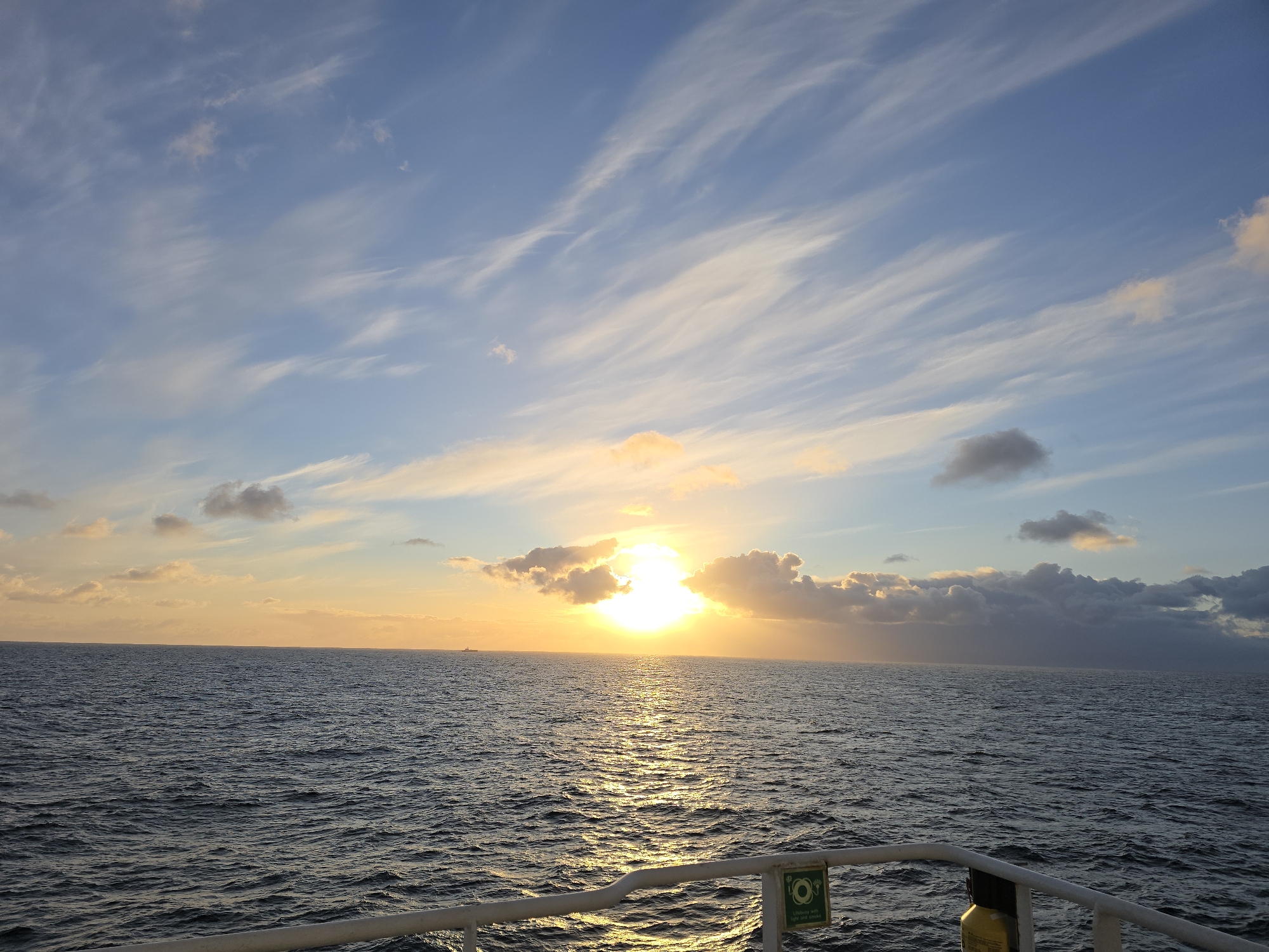 The image shows a sunset over the ocean from the perspective of a boat or ship. The sun is near the horizon, partially obscured by clouds. The sky is a mix of blue and wispy white clouds, with orange and yellow hues near the sun. The ocean is dark with bright sunlit reflections. A railing of the boat is visible in the foreground. A green sign with a white life preserver symbol is on the railing. The location is an unidentified area, at sea.