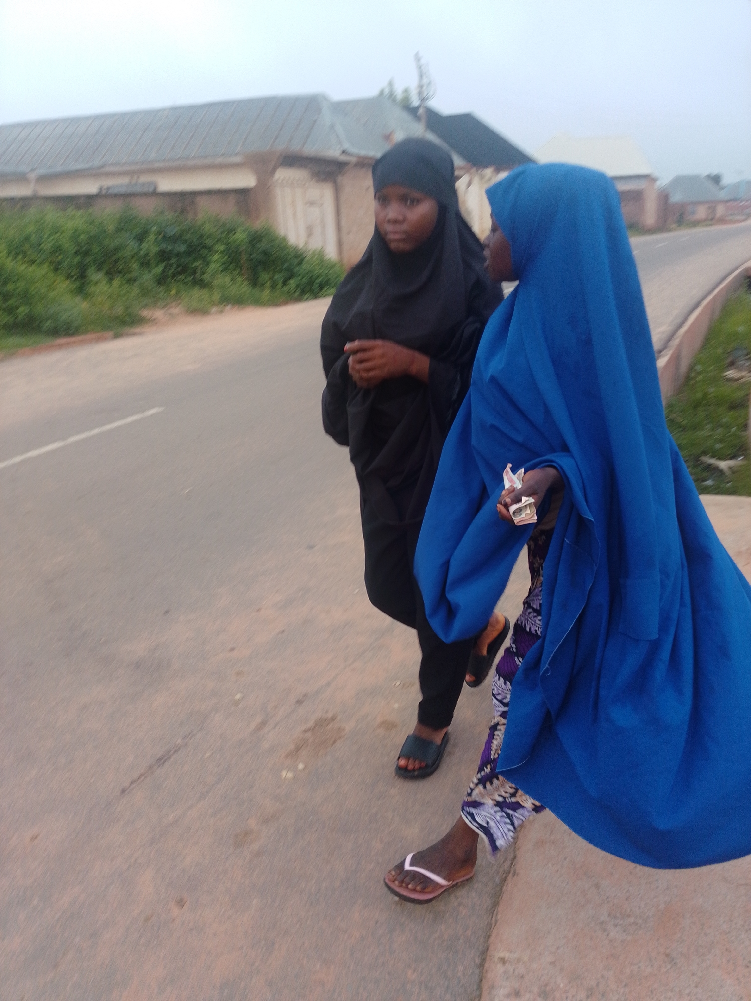 Two young women, dressed in Islamic attire, walk on a paved road. The woman on the left wears a black hijab and abaya, while the woman on the right is adorned in a vibrant blue hijab and a patterned skirt. The woman in blue is holding several banknotes in her hand. They are walking alongside a roadside, with a row of buildings and lush greenery in the background. The scene appears to be in a moderately populated area, possibly a town or a suburban district, under an overcast sky.