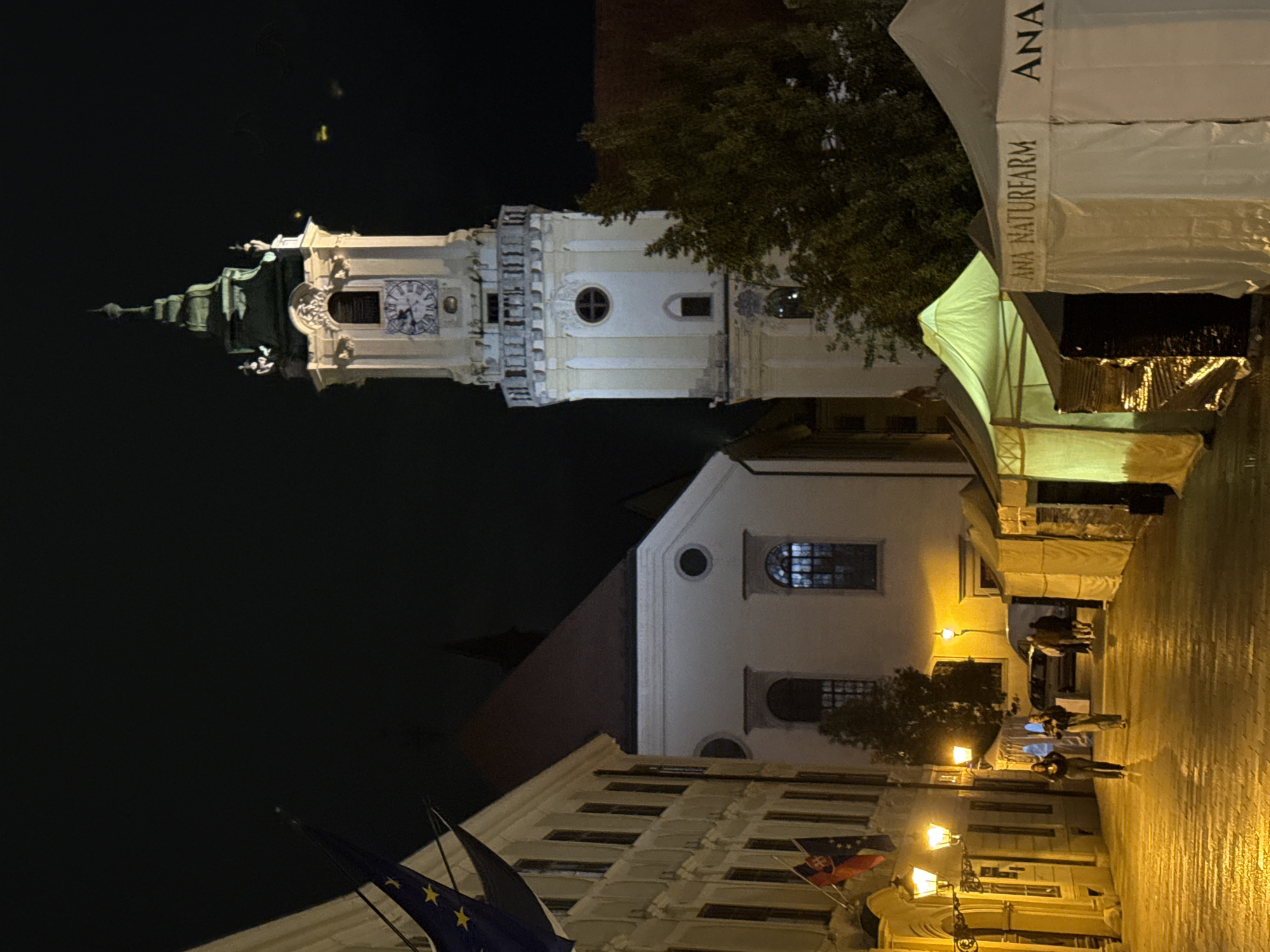This image depicts a street scene at night in Bratislava, Slovakia, specifically on Michalská Street, near Michael's Gate. A prominent clock tower, likely part of the Old Town Hall, stands tall against the dark sky, its clock face illuminated. In the foreground, several market tents are set up, one with the text "ANA NATURFARM" visible. Two people are walking down the cobblestone street, illuminated by streetlights and the glow from the tents. The street is wet, suggesting recent rain. European Union flags and a Slovak flag are visible on a building to the left, adding to the sense of place. The overall atmosphere is that of a quiet evening in a historic urban setting.