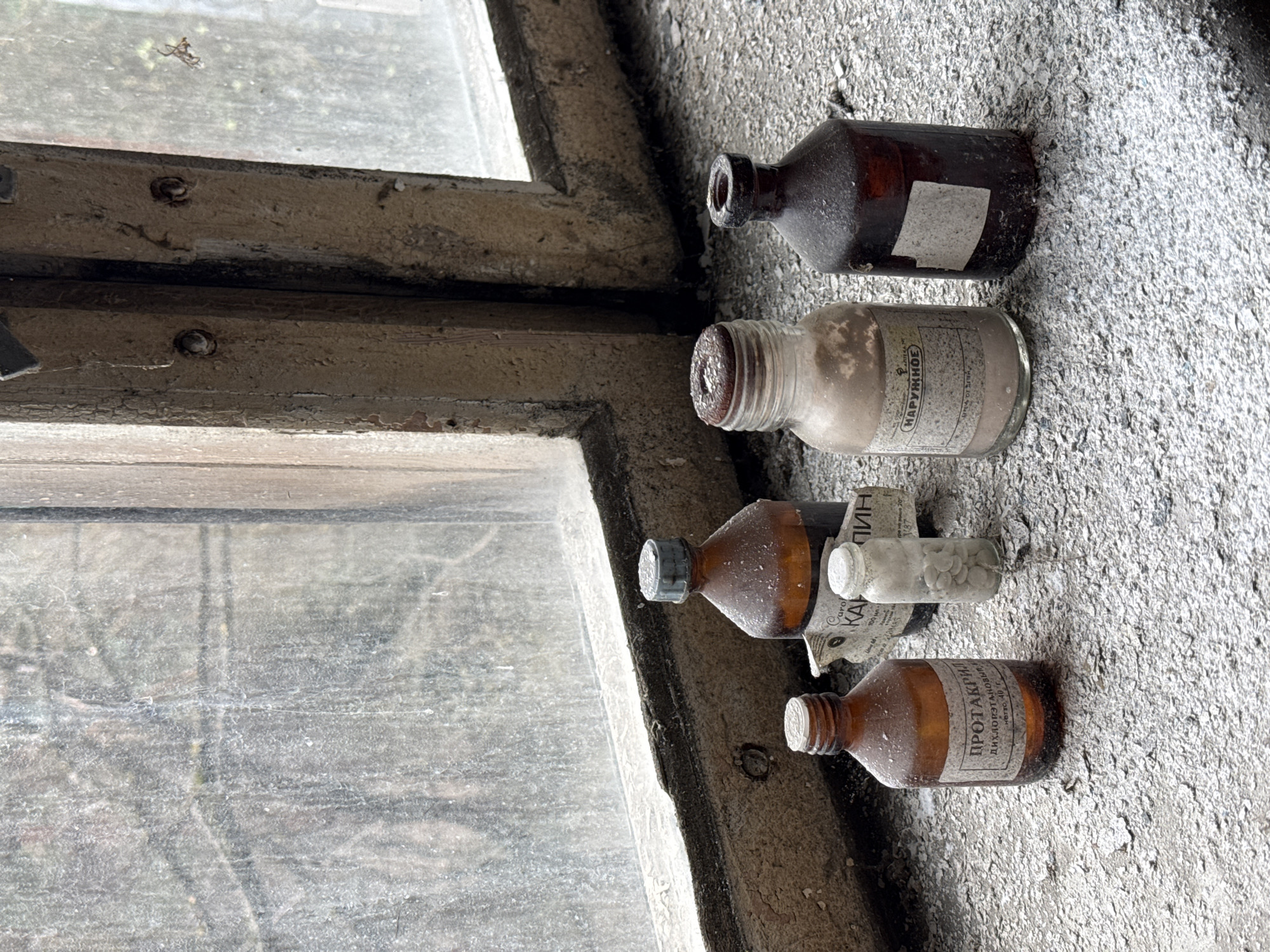 The scene depicts a window ledge in a dilapidated building, likely in Prypiat, Ukraine, based on the presence of Cyrillic text on the medicine bottles. Four glass medicine bottles are resting on the concrete ledge. The first bottle is brown and unlabeled. The second bottle is clear with a label that includes the word "HAPYЖHOE" in Cyrillic. The third bottle is brown and has a label with Cyrillic text, a partially torn label from another item, and a small clear bottle filled with white pills resting on top of the brown bottle. The fourth bottle is also brown and its label says "ПРОТАКРИЛ" in Cyrillic. The window is dirty and the frame is peeling. The concrete around the bottles is crumbling and covered in dust and debris.