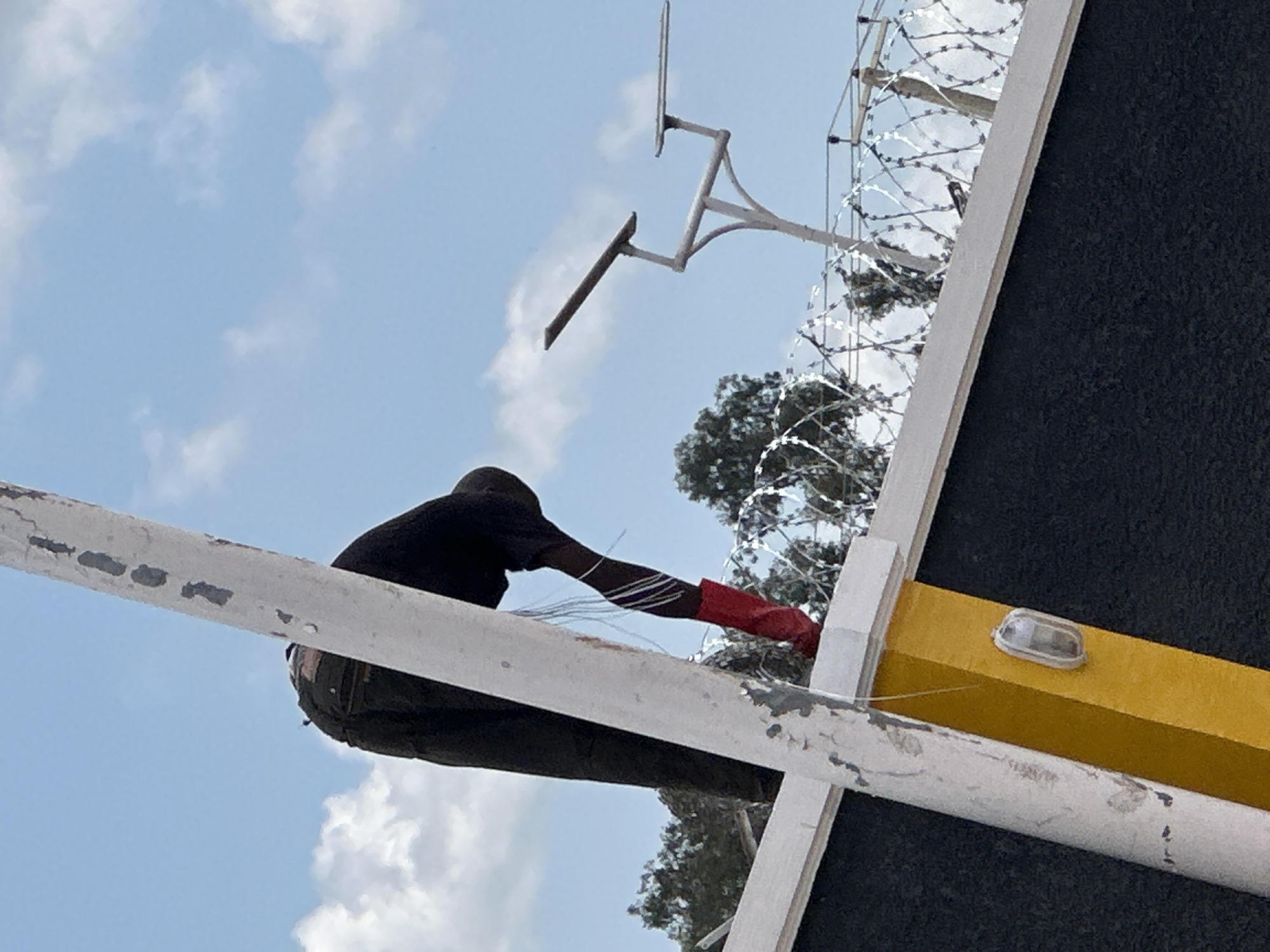 A person, wearing dark pants and red boots, is crouched on top of a dark-colored wall topped with barbed wire. The person appears to be reaching down or adjusting something near their feet. A white pole with yellow accents runs diagonally across the foreground. In the background, a bright blue sky with scattered clouds is visible, along with trees and a modern street lamp. The location is identified as Barakin Lerawi, Nigeria.