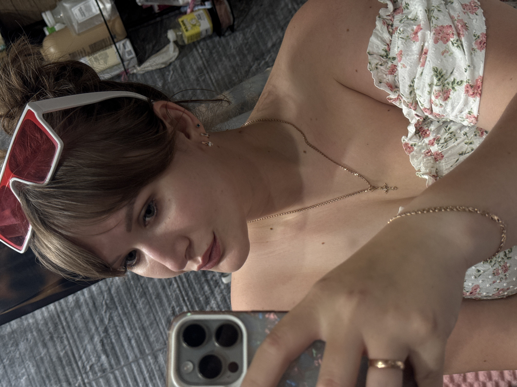 This media file is a portrait of a young woman, taken from the shoulders up. She is indoors, lying down on a gray surface that appears to be a bed or couch. The lighting suggests it could be daytime, but the exact time is not clear.
The woman has her hair pulled up in a messy bun and is wearing a white off-the-shoulder top with a floral pattern of pink roses and green leaves. She has on large, white-framed sunglasses with red lenses, which are pushed up onto her head. Around her neck, she wears a delicate gold chain with a small cross pendant. On her wrist, she has a thin gold chain bracelet, and on her right ring finger, a simple gold band. Her left ear has two small piercings, one with a blue stone and another with a clear gem.
She is holding a smartphone in her right hand, angled as if taking a selfie. The phone case has a reflective, multi-colored pattern. Her expression is relaxed, with a slight smile and her gaze directed towards the phone.
In the background, out of focus, there are various objects on what appears to be a shelf or surface. These include several bottles and containers, one of which is a yellow bottle with some text that is not legible. There are also some white cloths or papers. The overall setting seems casual and personal. There is no clear indication of the weather or a specific event taking place, beyond the act of taking a selfie.