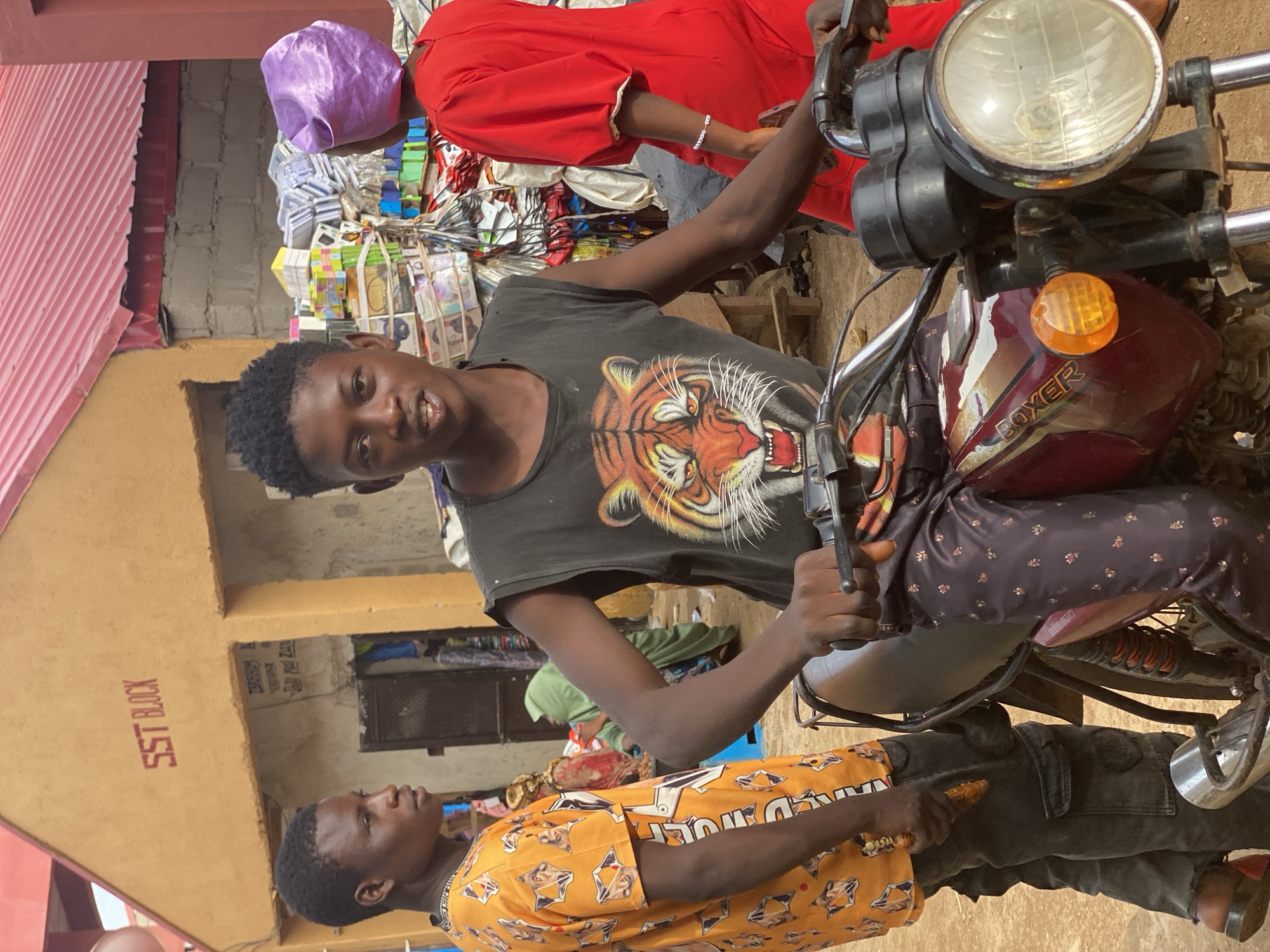 The image, taken in what appears to be the city of Kafanchan B, Nigeria, depicts a young man sitting on a red Boxer motorcycle. He wears a dark sleeveless shirt with a tiger print. Another individual stands to his left, wearing a yellow shirt with patterns, appearing to be holding an object. The backdrop includes a building labeled "SST BLOCK" and a stall or shop stocked with various items. Several other people are present in the background, one wearing a red garment and another wearing a purple head covering. The lighting suggests it is daytime.