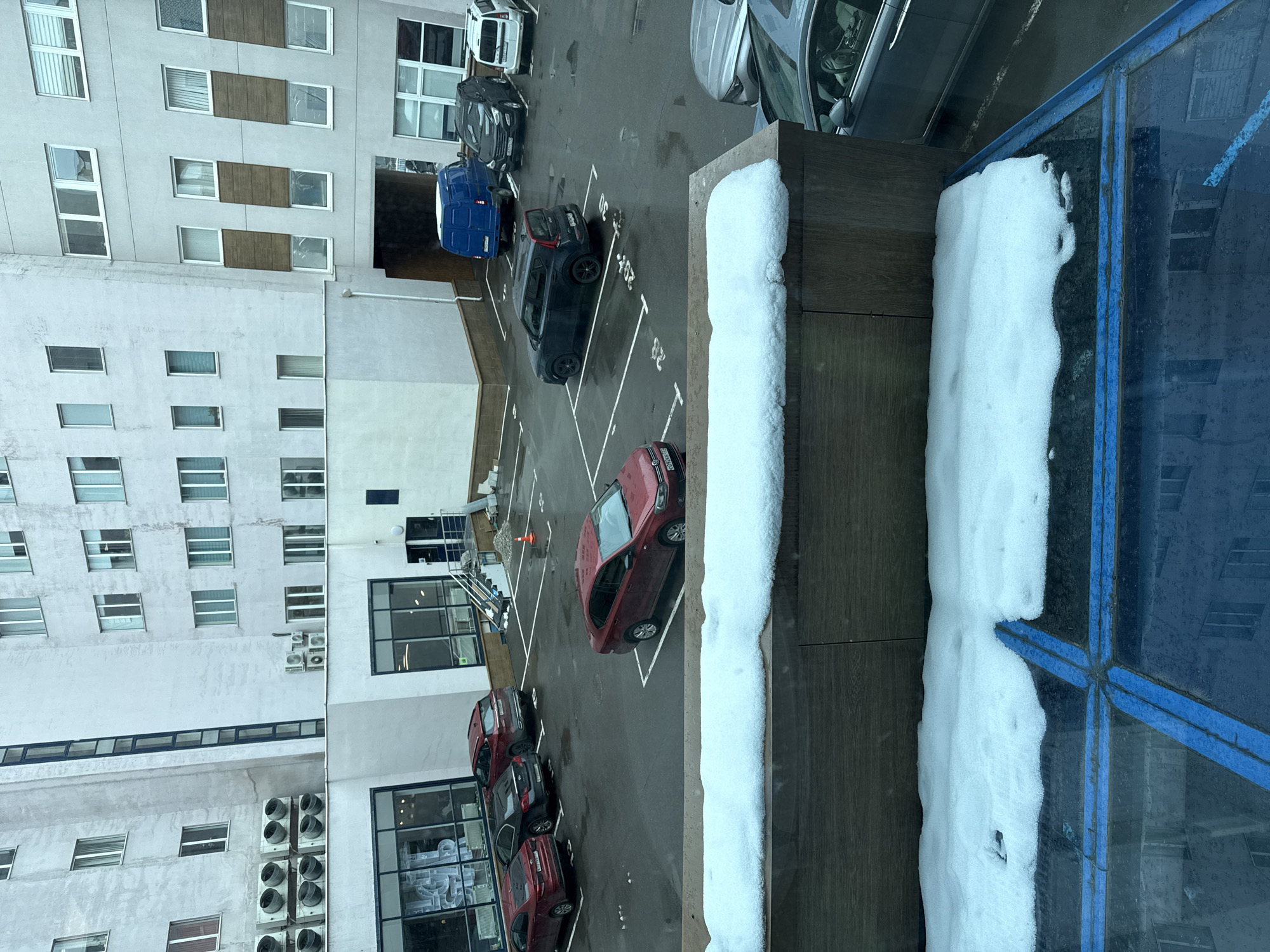 The image captures an elevated view of an outdoor parking lot surrounded by modern, multi-story buildings, likely in an urban or suburban setting like Moscow. The scene is set during the day, under overcast skies, and reflects cold, winter conditions.
In the immediate foreground, a window ledge covered with a thick layer of pristine white snow extends across the bottom, partially obscuring a blue-framed glass structure on the right side, which also bears snow accumulation.
The central part of the image features an asphalt parking lot with clearly marked white lines indicating individual parking spaces. Numerous cars are parked, including a prominent red hatchback/sedan in the closest space with a visible Russian license plate (starting with "M938"). Further back, other vehicles include a dark grey/black car, a blue van, another black car, a white car, and a silver sedan. A small orange cone is visible further back in the parking area.
The background is dominated by the facades of the buildings. On the left, a light-colored (possibly white or light grey) building features multiple rectangular windows, some of which are flanked by dark wooden-like panels. Several external air conditioning units are mounted on its facade. Another building is partially visible on the right, featuring large windows and similar modern architectural elements.
No people are visible in the scene. The parking lot appears damp, suggesting melting snow or recent precipitation, but is not heavily snow-covered. Visible text includes parking space numbers "30" and "32" painted on the asphalt.