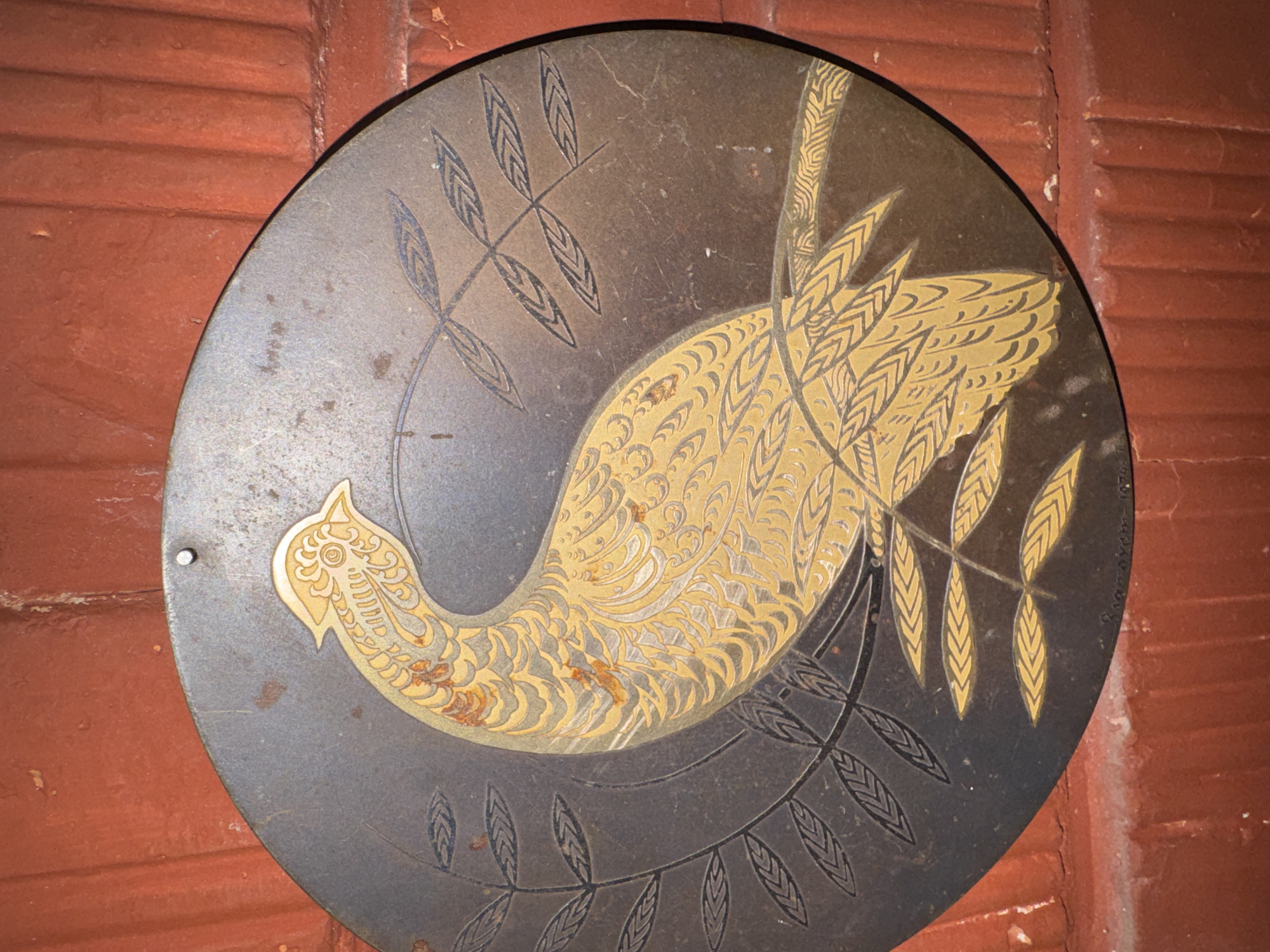 The image shows a circular metal plaque with a relief of a bird, likely a pheasant, rendered in gold and silver tones against a dark, textured background. The bird is depicted in profile, with its head turned upwards and its tail fanned out. Stylized leaves and branches surround the bird. The plaque has a small hole near the top, suggesting it is meant to be hung. A faint inscription is visible along the bottom edge of the plaque. The plaque is mounted on a red, brick-like surface. The city of Таврово, Russia is mentioned as the location.