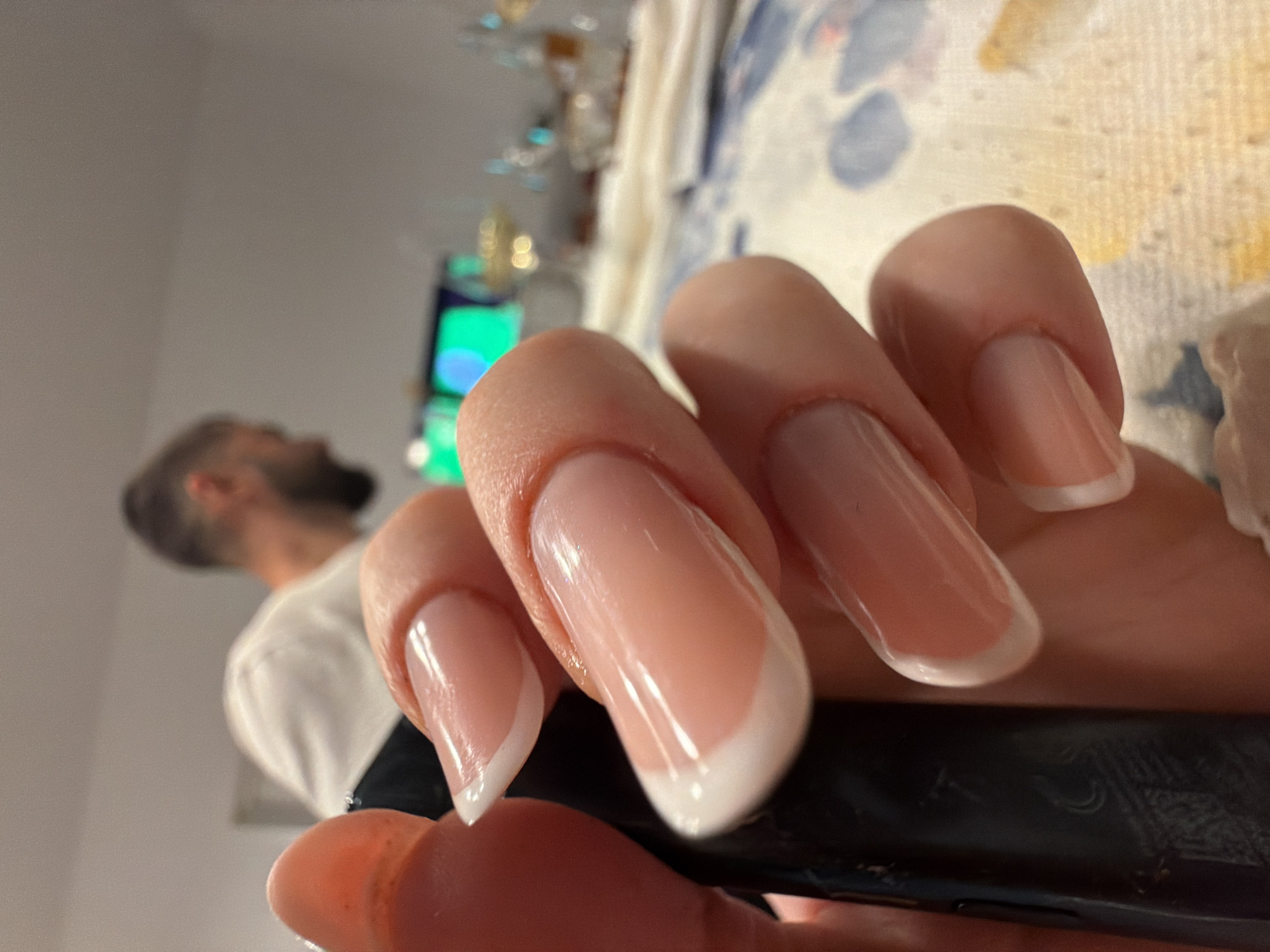 The image is a close-up of a person's hands with french manicured nails, holding a black cylindrical object. In the blurred background, a man with short dark hair and a beard is visible, wearing a white t-shirt. He appears to be in an indoor setting, possibly a home, with a table in the foreground that has a patterned cloth and some glassware. There is a screen displaying colorful lights in the far background. The scene does not provide any specific visual cues to place it in Florești, Romania.