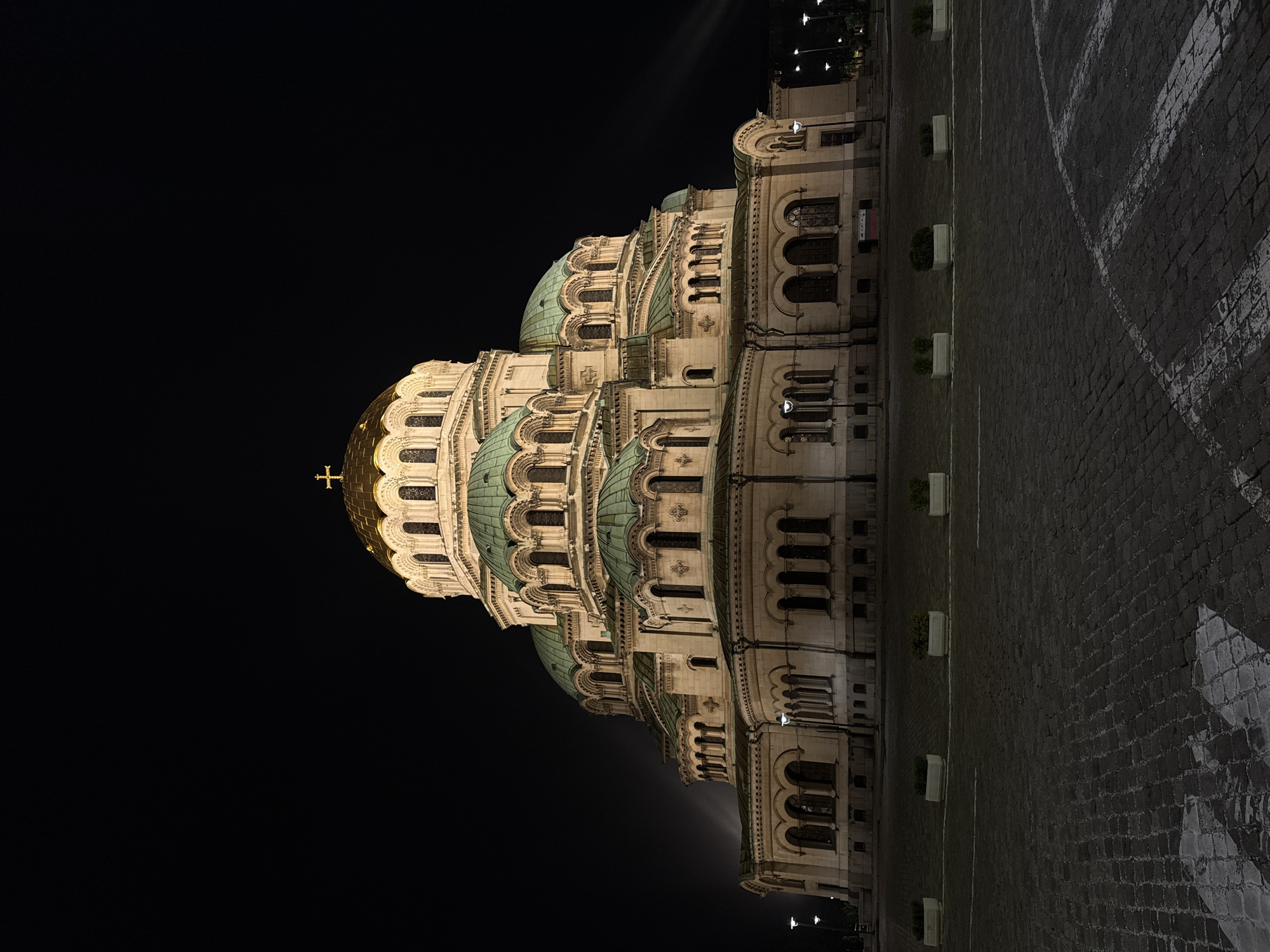 The image shows the St. Alexander Nevsky Cathedral in Sofia, Bulgaria, at night. The cathedral's illuminated facade stands out against the dark sky, showcasing its architectural details, including domes, arches, and windows. A golden dome topped with a cross is prominently visible. The building stands next to a cobblestone ground with white stripes. The surrounding area appears dark, with limited ambient lighting.