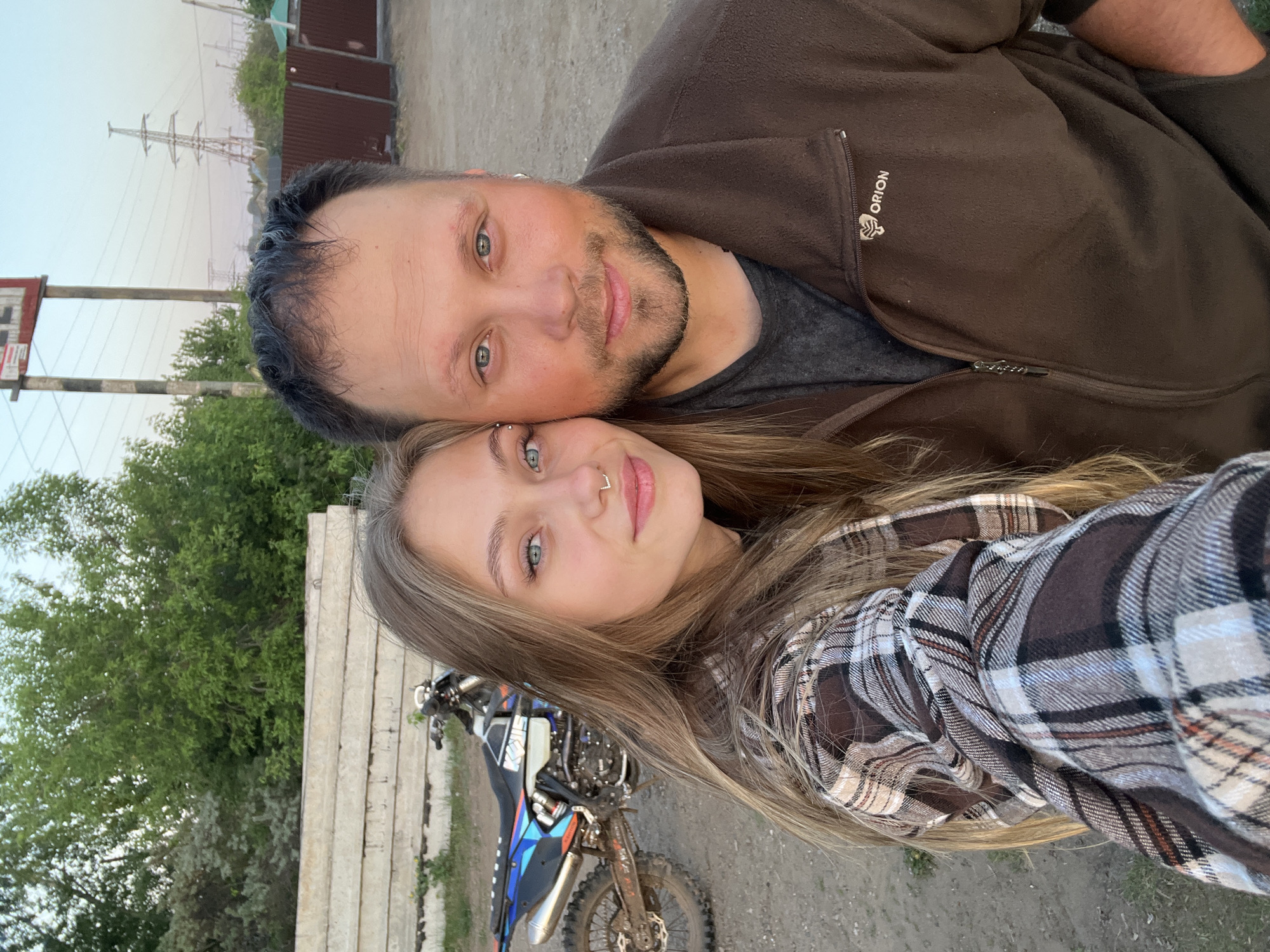 Two individuals are posing for a selfie outdoors. The male is wearing a brown zip-up jacket with the word "ORION" visible on the upper left. The female, positioned lower in the frame, is wearing a plaid flannel shirt. A dirt bike is visible in the foreground. The background includes vegetation, a concrete wall, a metal building, and power lines. The location is the city of Chaikovsky, Russia.