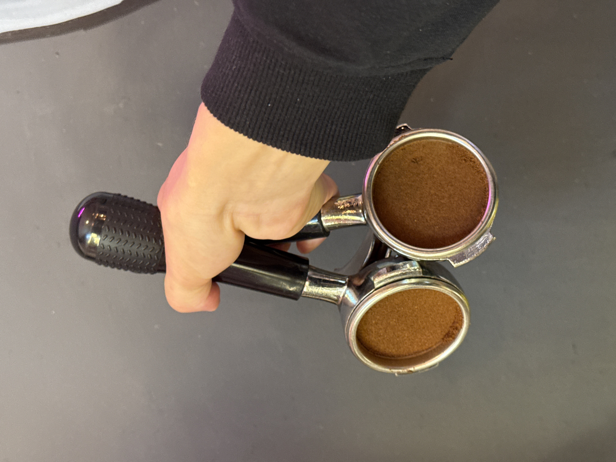 A close-up shot shows a hand holding a black-handled portafilter with two baskets filled with ground coffee. The portafilter is held horizontally, with the baskets facing downwards. The hand is wearing a black long-sleeved garment on the wrist. The background is a plain dark gray wall. The image was taken in Moscow, Russia.