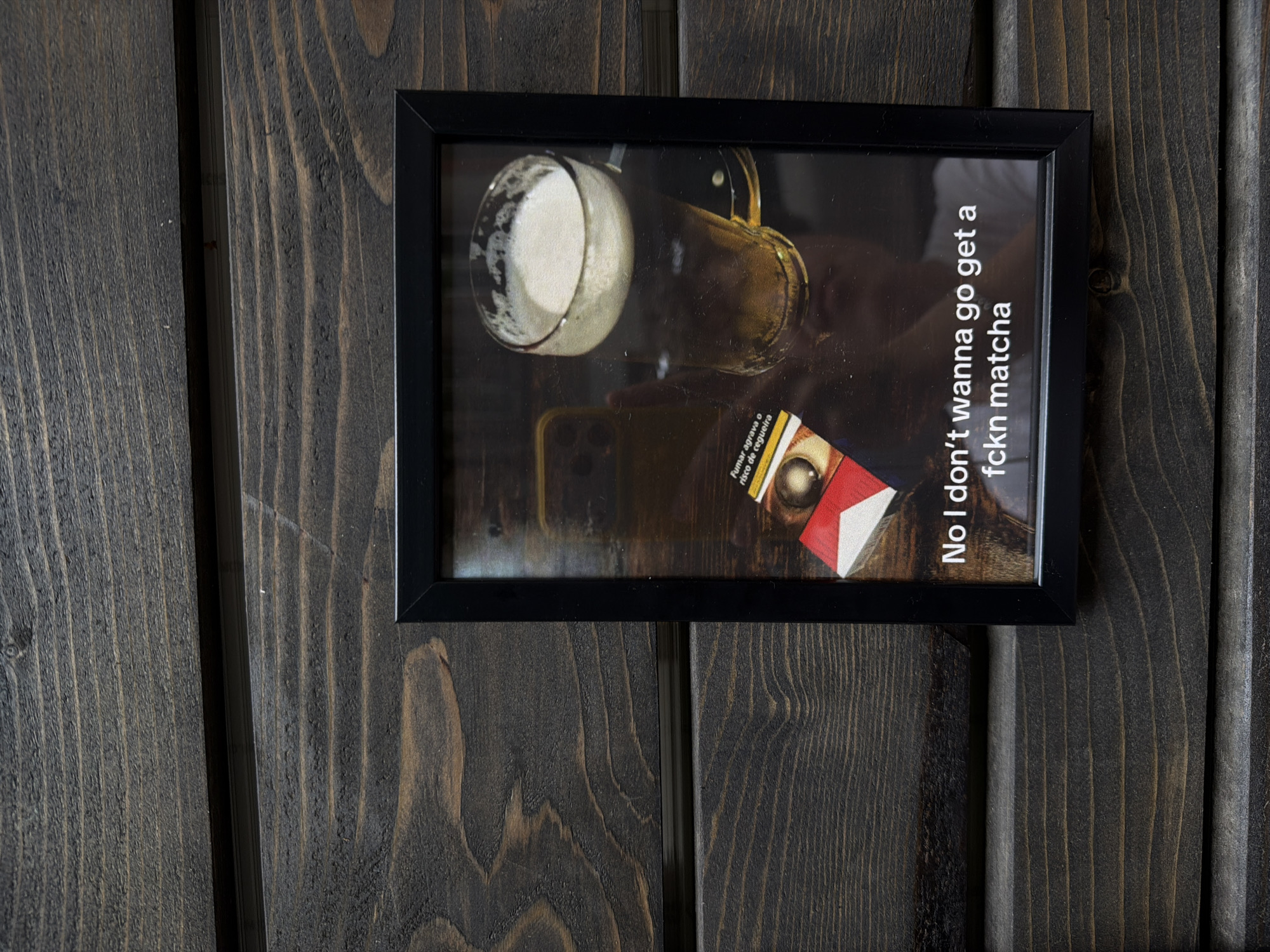 A framed print hangs on a dark wooden panel wall. The print features an image with a glass of liquid, a gold object, and a cigarette pack. White text on the right side of the print reads, "No I don't wanna go get a fckn matcha." The cigarette pack has the text "Fumar agrava o risco de cegueira." This indicates that the picture originates from a Portuguese-speaking country. The location appears to be an interior space in Belgrade, Serbia.