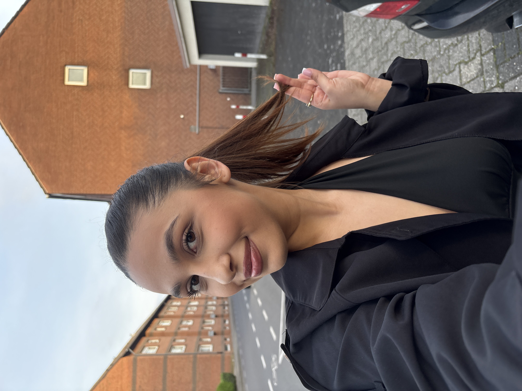 The image depicts a woman in Mönchengladbach, Germany. She is smiling at the camera and holding a strand of her hair. She is wearing a black top and jacket. Behind her is a brick building with windows. A road and part of a car are also visible.