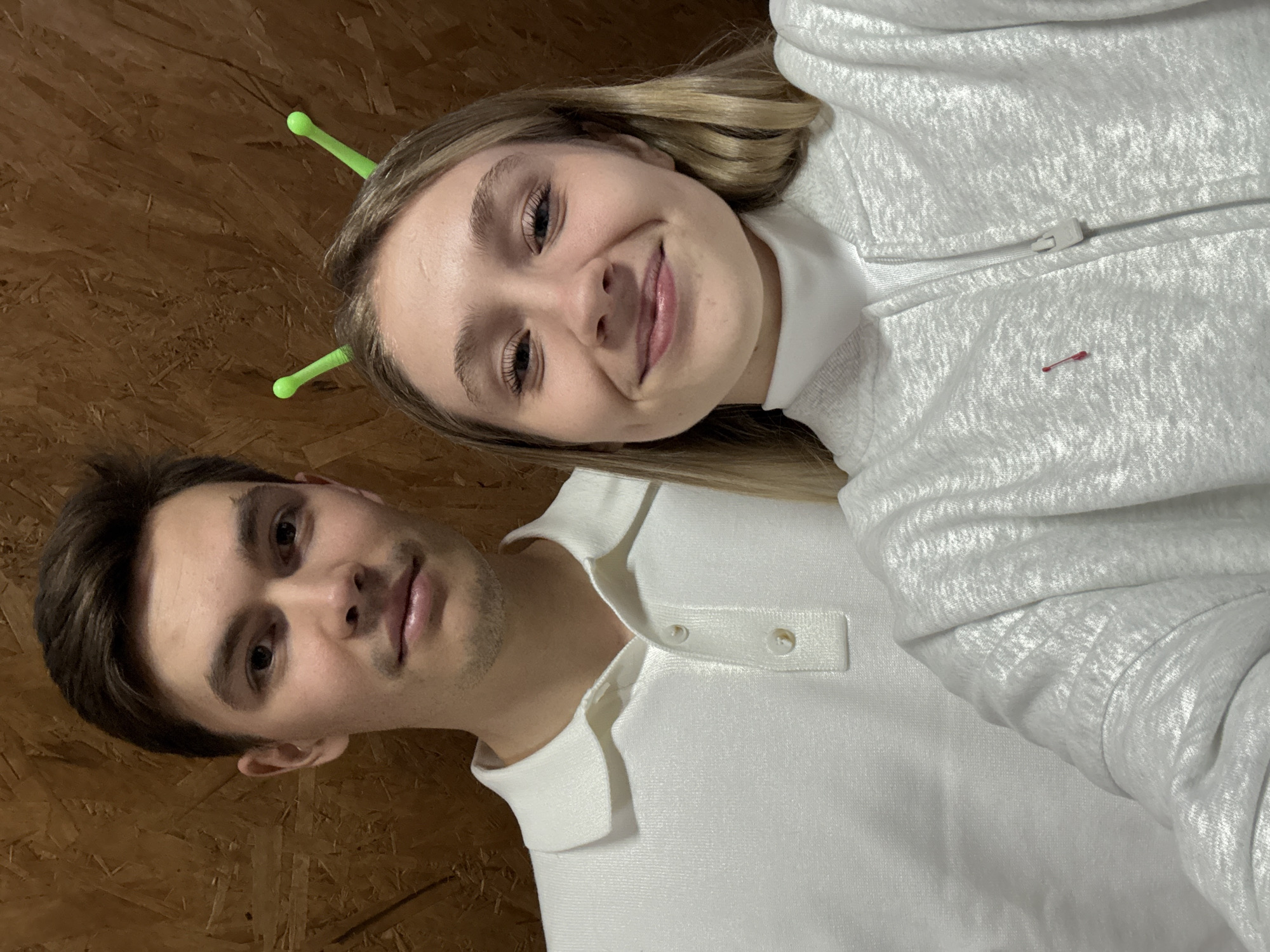 The image shows a selfie of two people indoors, possibly against a plywood wall. The individuals are both wearing white tops; the woman is wearing a light grey hoodie with a red safety pin, and the man is wearing a white polo shirt. The woman also has two green objects in her hair, possibly hair accessories. The location is described as being in the city of Прикамский, Russia.