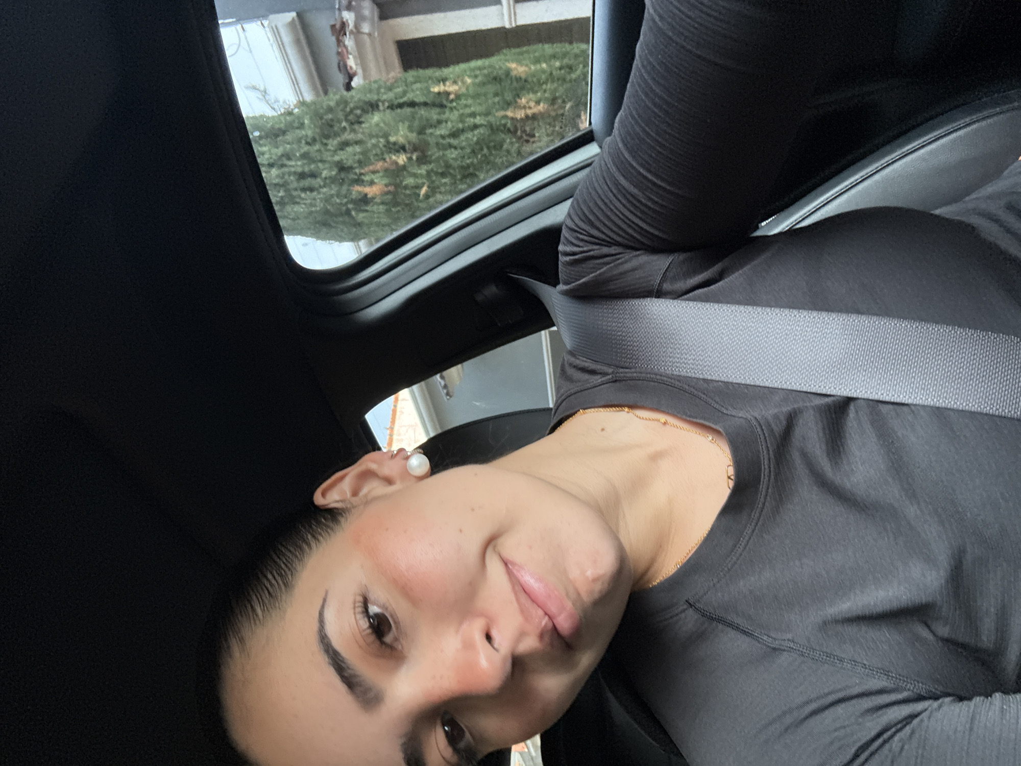 A selfie shows a woman wearing a pearl earring, a gold necklace, and a black long-sleeved shirt, buckled into a car seat with a gray seatbelt. The car's sunroof is open, showing greenery and a building outside. The location is San Bernardino Tlaxcalancingo, Mexico.