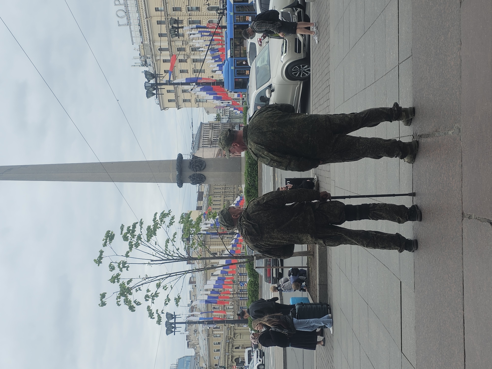 The image shows two men in military fatigues standing on a sidewalk in Saint Petersburg, Russia. They are near the Alexander Column in Palace Square. The men appear elderly, with one using a walking stick. The background shows buildings adorned with numerous Russian flags. Other pedestrians and vehicles are present in the vicinity. The sky is overcast.