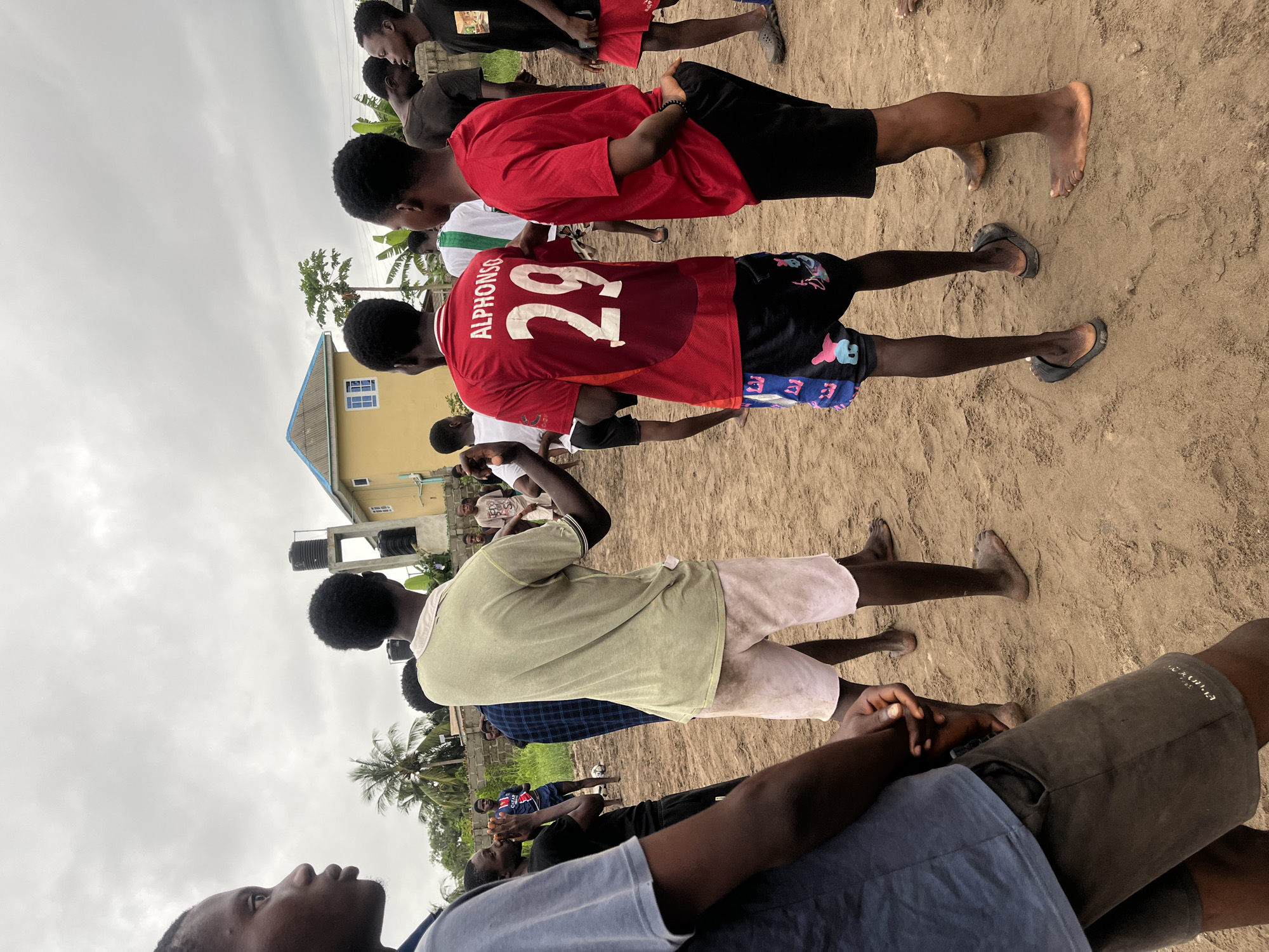 A group of young men stand on a dirt field in Badagry, Nigeria, with a yellow building and water tanks in the background. The sky is overcast. Two of the men in the foreground are wearing red t-shirts with the number "29" and "Alphonso" printed on the back of one. One man wears light-colored shorts, while another wears black shorts. Other men are dressed in various casual clothing. A soccer ball is visible in the background. The scene appears to be a casual gathering or practice.