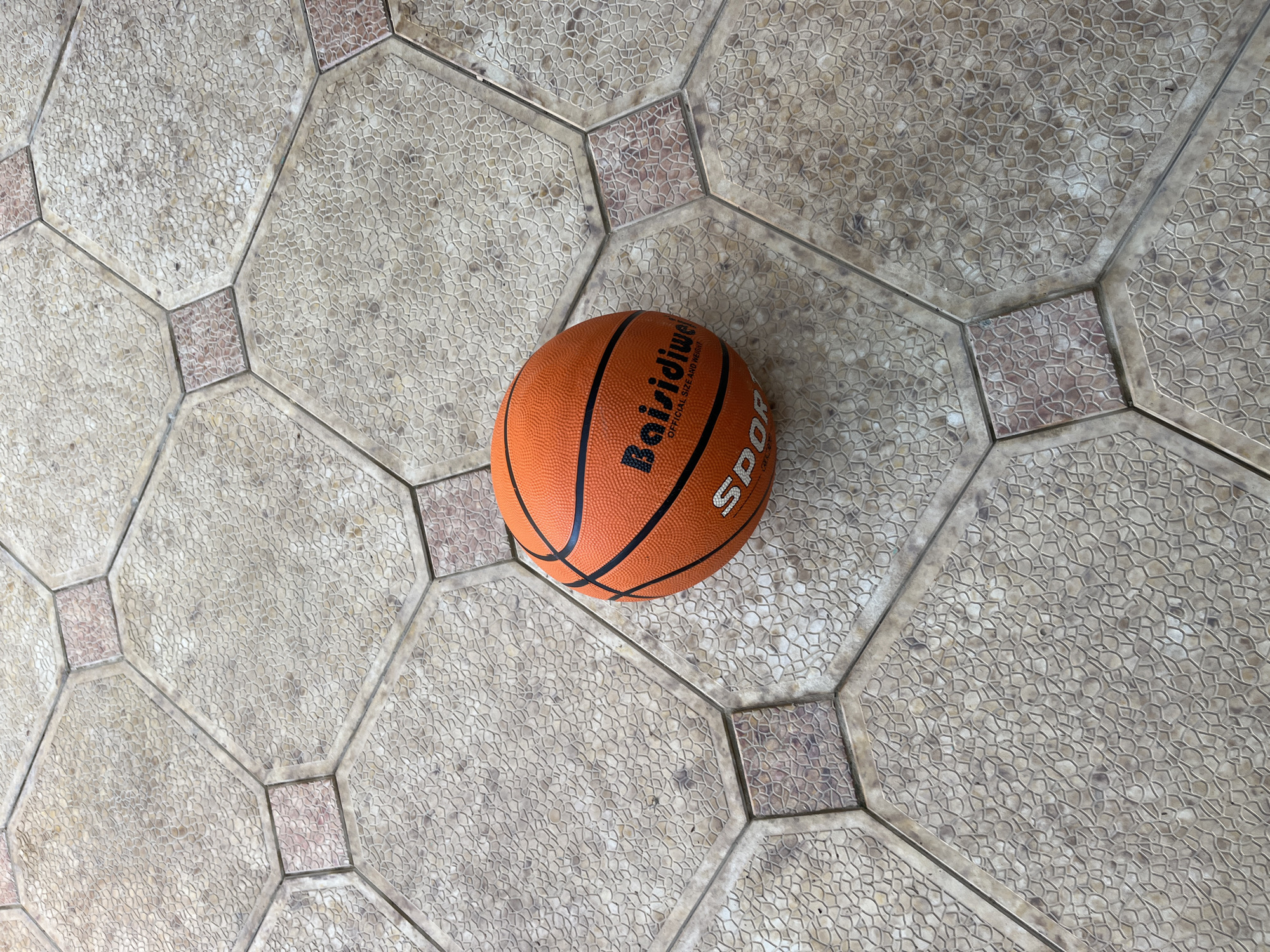 An orange basketball rests on a tiled surface composed of large, light-beige octagonal tiles connected by smaller, square, reddish-brown tiles. The tiles feature a textured surface resembling small pebbles. The word "Baisidiwe" and the text "OFFICIAL SIZE AND WEIGHT" are printed on the basketball. The term "SPORT" is also visible on the ball. The setting may be located in Kaspiysk, Russia, given the query context, although that cannot be definitively confirmed from the image alone.