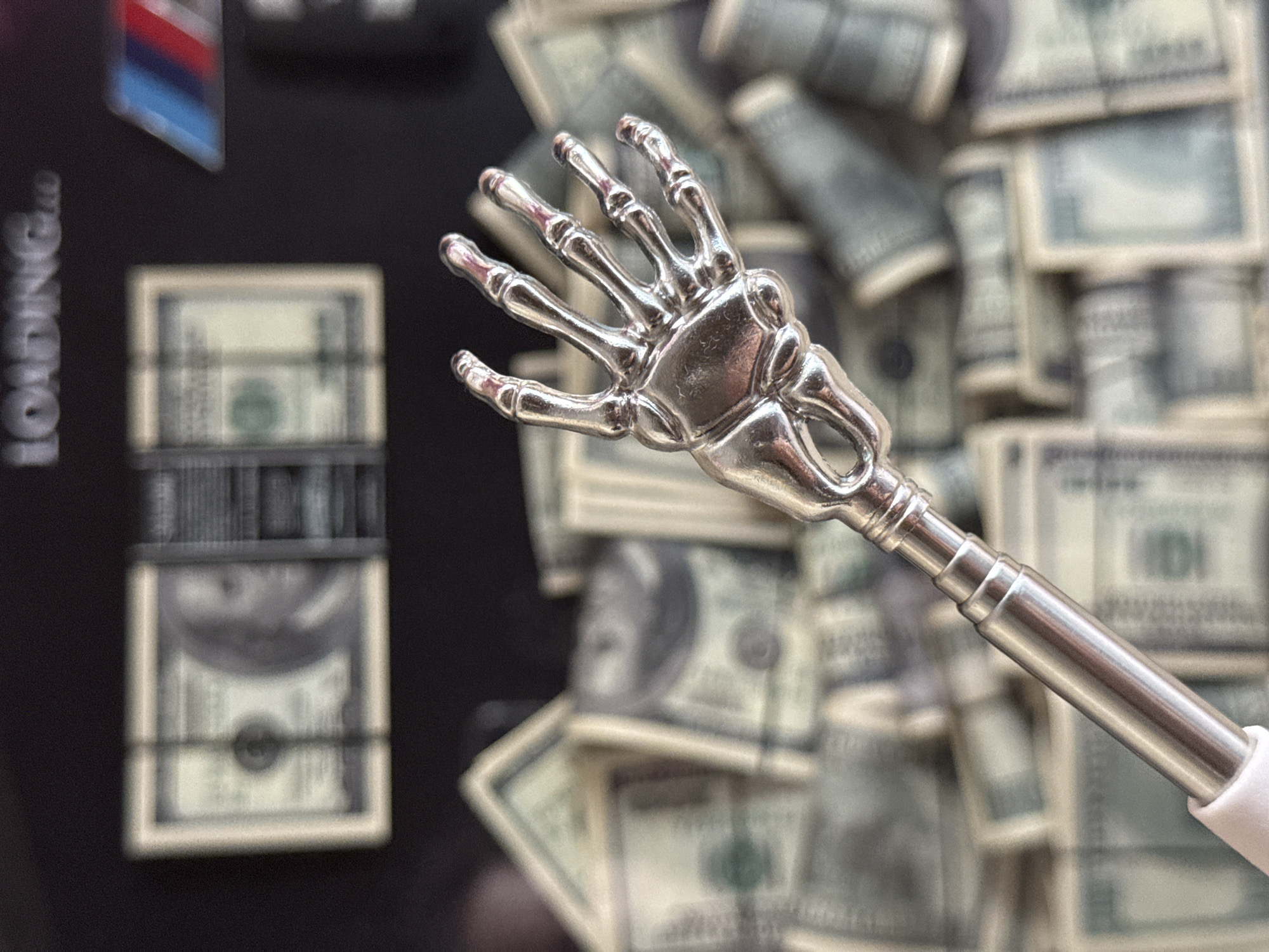The image displays a close-up shot of a metallic skeleton hand attached to a telescoping rod. The hand is positioned in the foreground, with its fingers extended. Behind the hand, a pile of US hundred-dollar bills is visible, some stacked neatly and others scattered. In the upper background, there is text that appears to read "LOADING..." and a blurred image of a BMW logo. The scene is dimly lit, with the metallic hand reflecting the surrounding light. The image does not provide any information or context related to the city of Yekaterinburg, Russia.