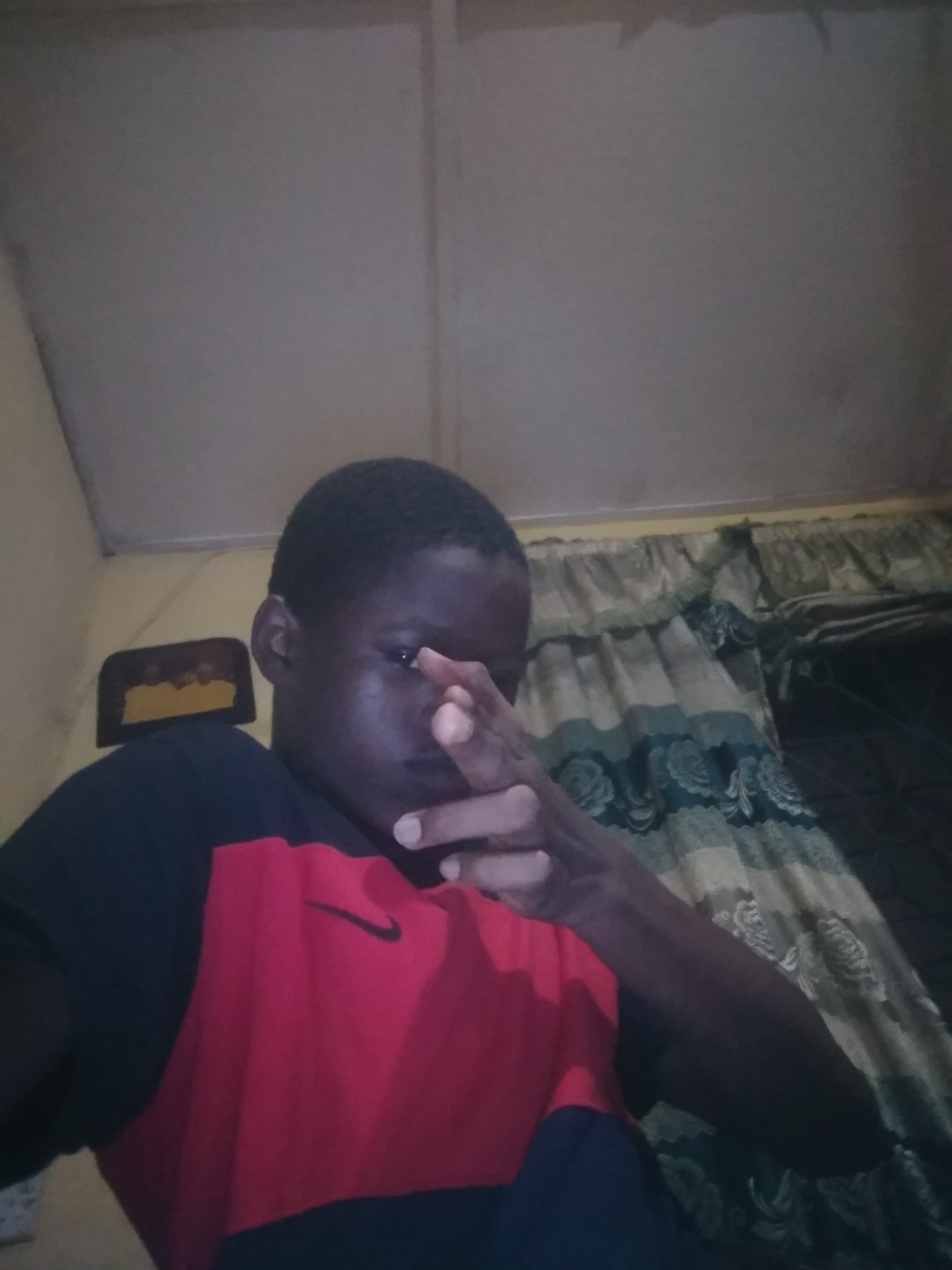A medium shot presents a young person indoors, likely in a domestic setting. The individual is wearing a shirt with a black Nike logo on the red panel. The background includes a curtain with a patterned design. The ceiling is plain and appears to be made of panels. A small, framed object is visible on the wall. The person is holding up their hand, partially obscuring their face. The lighting is dim. The location is unknown.
