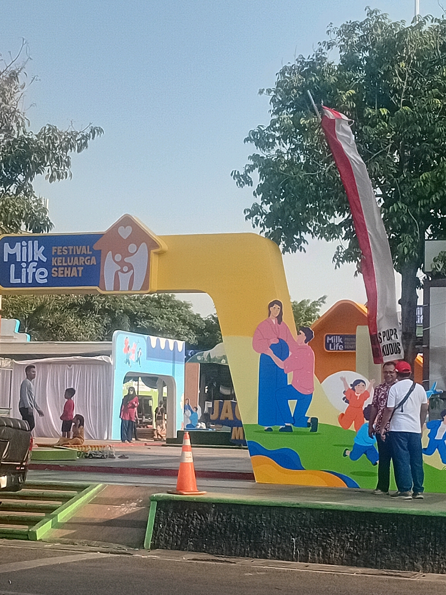 An outdoor festival is taking place in Kudus, Indonesia, with a large, colorful archway that reads "Milk Life Festival Keluarga Sehat" in English and Indonesian. The archway is adorned with illustrations of a family. A red and white Indonesian flag is visible, as is a tree. Several people are present, including a man in a gray shirt and a child in a red shirt on the left, and a woman in a pink hijab and a red top further back. Two men in the foreground are looking towards the archway, and a man in a white shirt and red cap is facing the camera. An orange traffic cone is positioned on the ground.